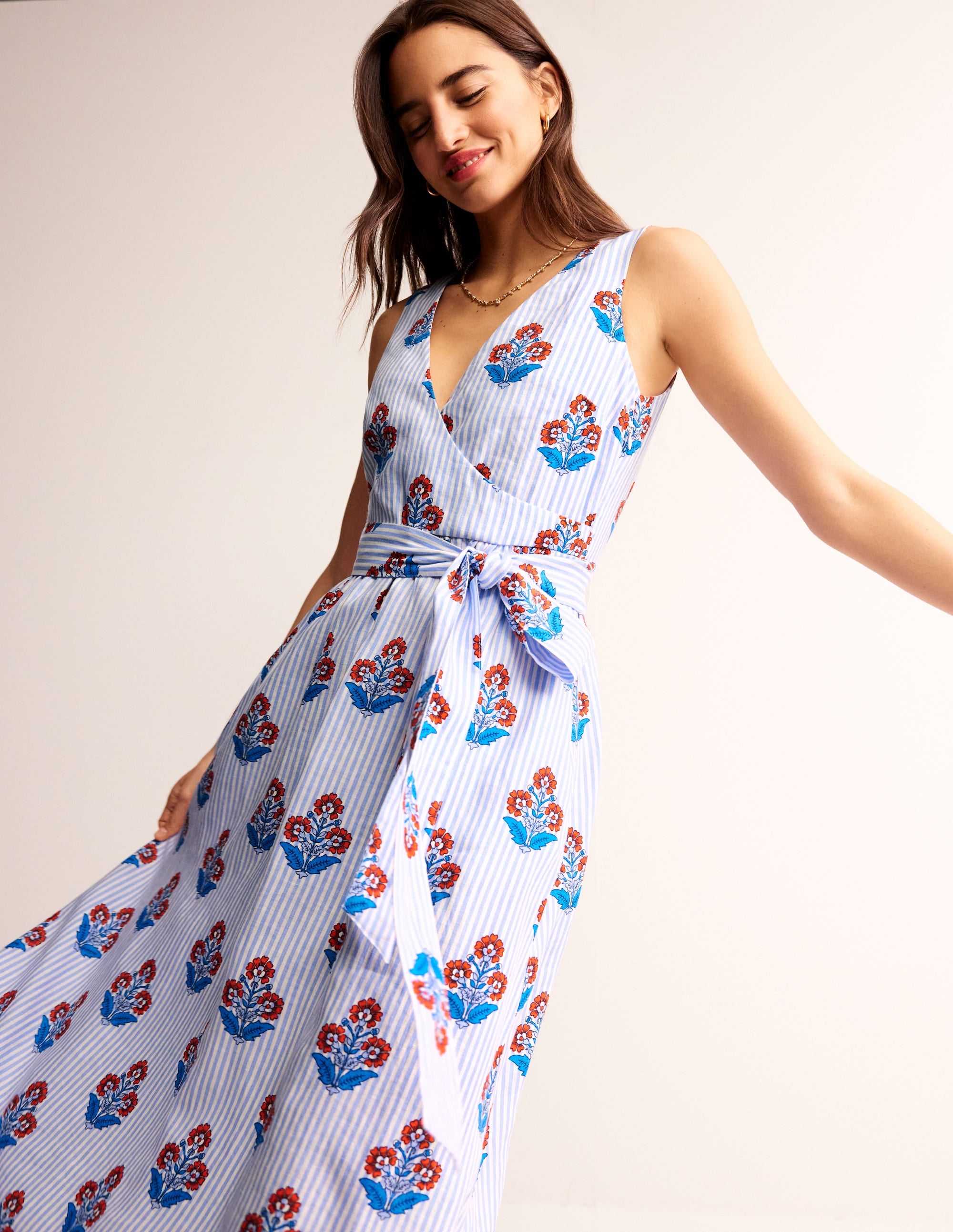 A woman wearing a mandarin orange linen wrap midi dress with a floral print, featuring a large bow at the front, sleeveless design, and a fitted waist.