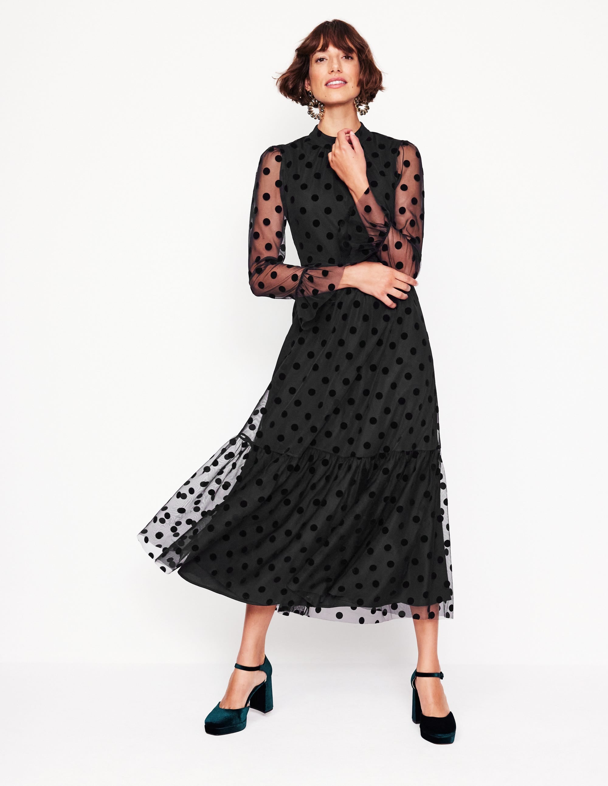 A woman wearing a black high neck tulle tea dress with sheer polka dot long sleeves, a large back bow, and a tiered skirt, paired with teal high heels.