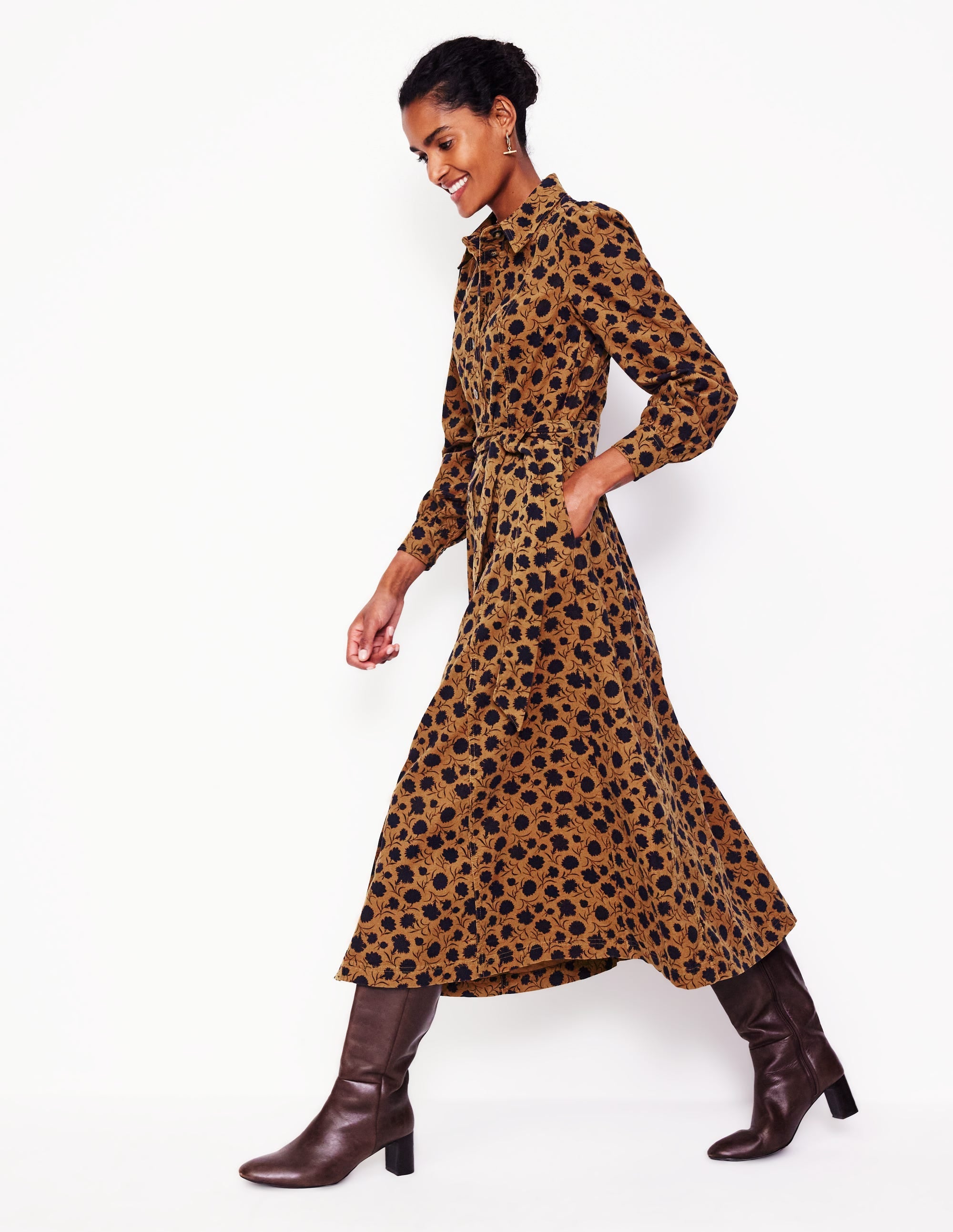 A woman wearing a brown and black floral corduroy midi shirt dress with a collar, long sleeves, a belt, pleats, and side pockets, paired with knee-high brown boots.