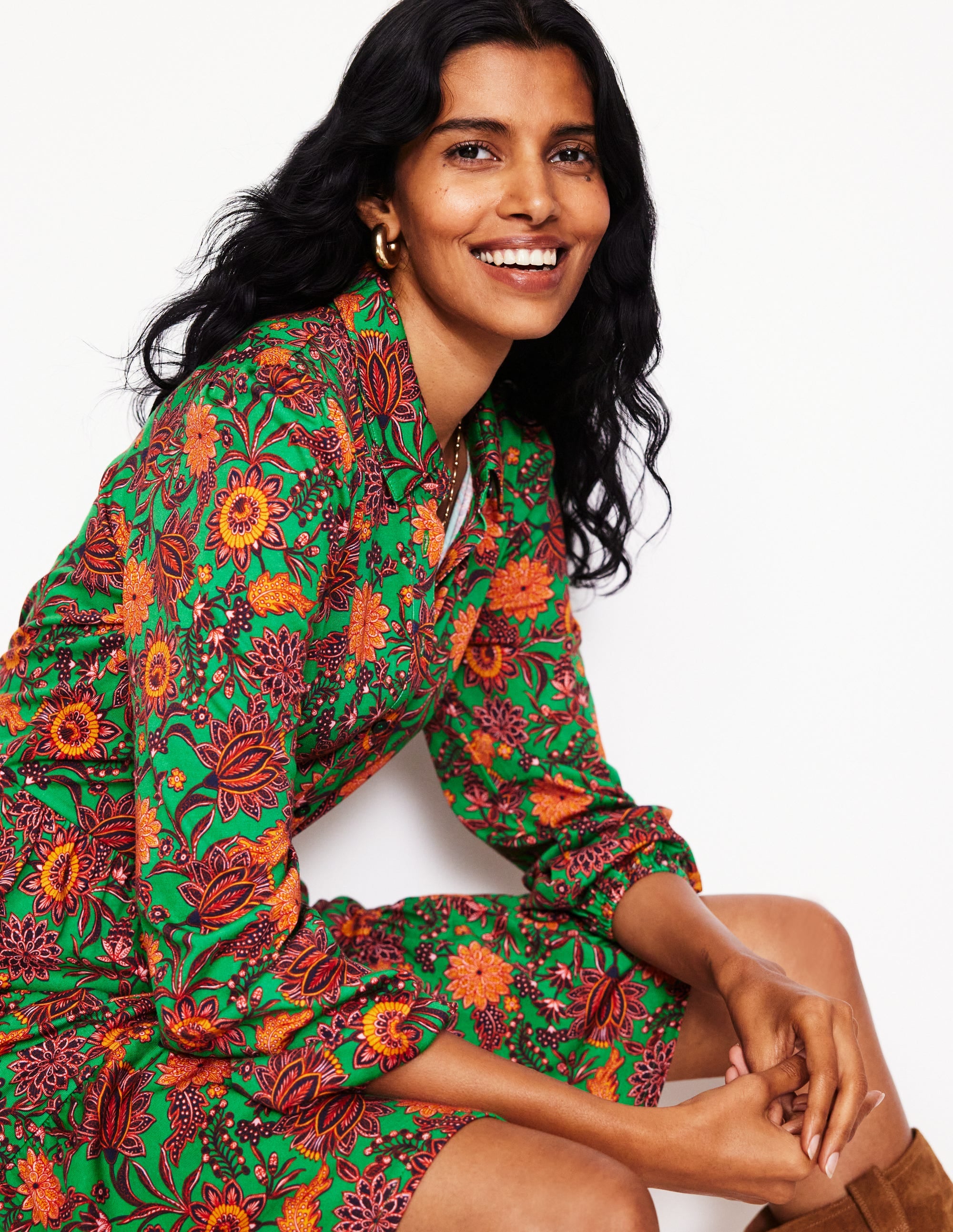 A woman wearing a vibrant green, tiered jersey dress with a floral pattern, featuring side pockets and a belt, sitting with her hands clasped.