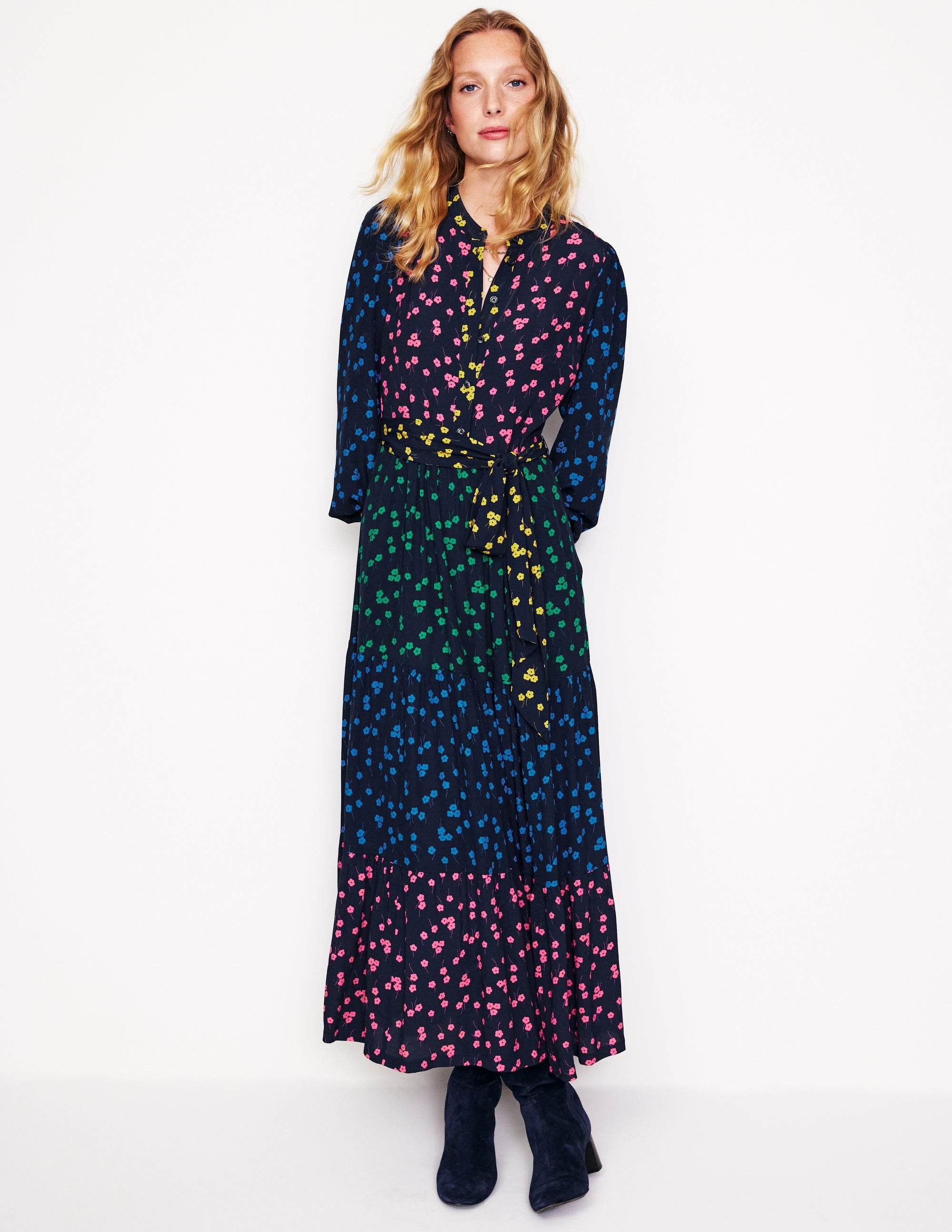 A woman wearing a long, colorful floral maxi dress with tiers, gathered sleeves, a matching belt, and side pockets.