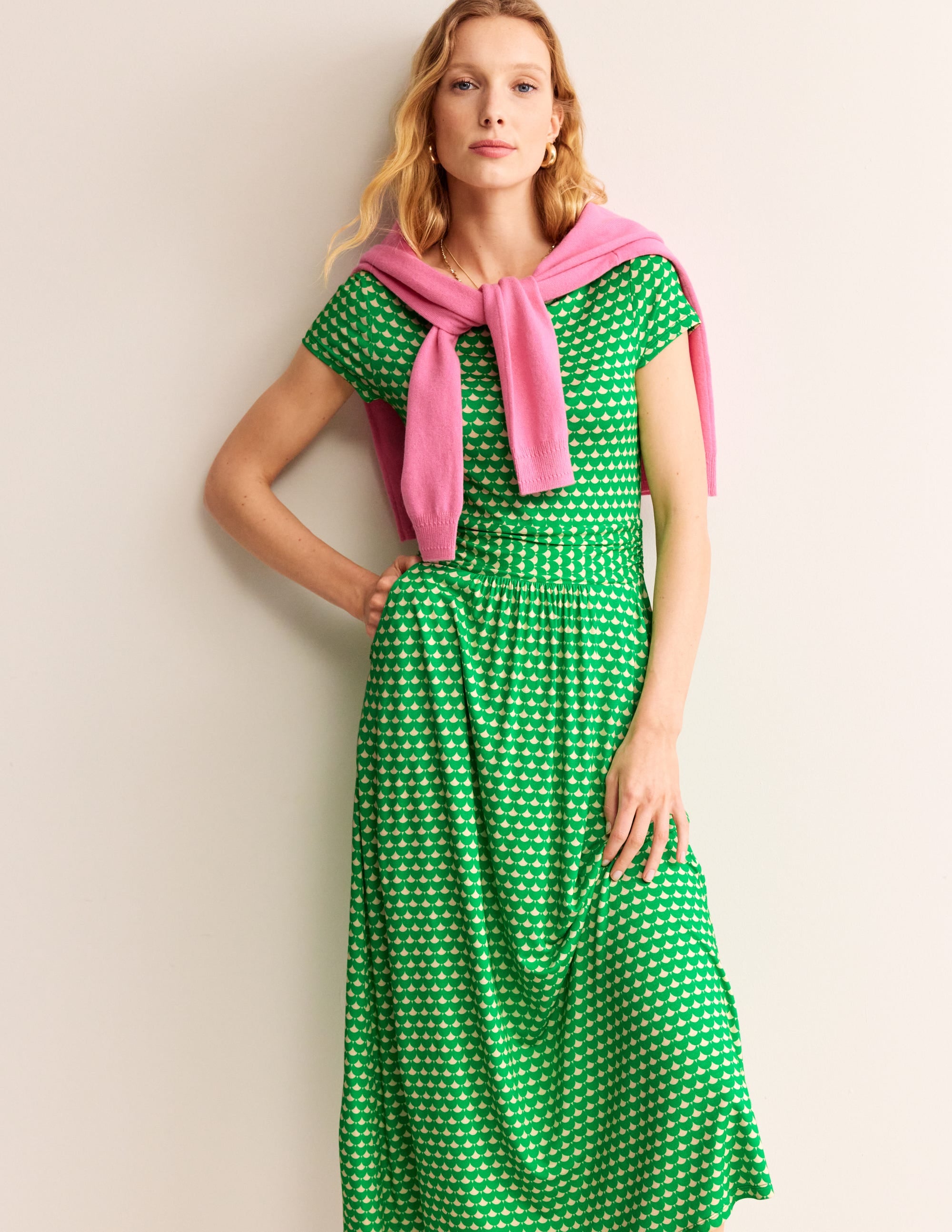 A woman wearing a Kelly Green midi dress with a crescent stamp pattern, fitted at the bust and waist, with a full skirt, side pockets, and a ruched waist.