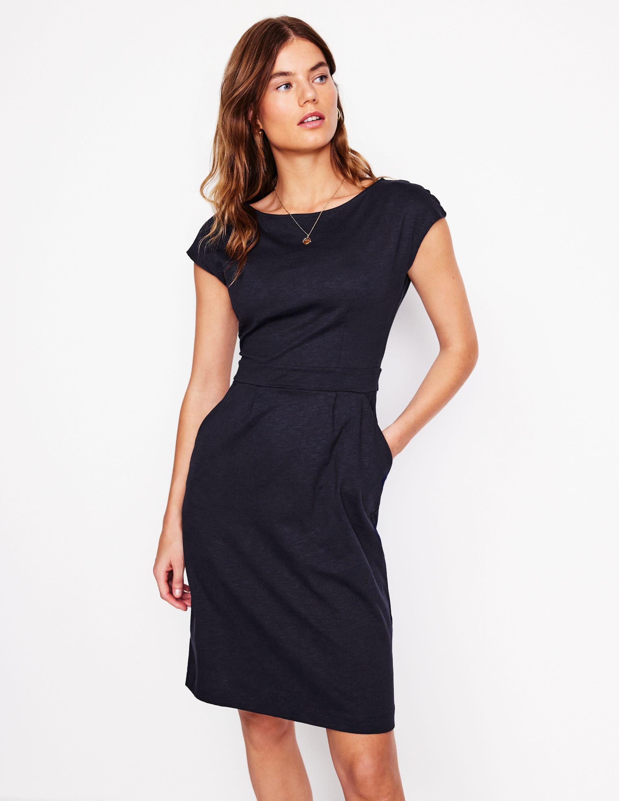 A woman wearing a navy jersey dress with a fitted waist, front pleat, and side pockets, styled casually.
