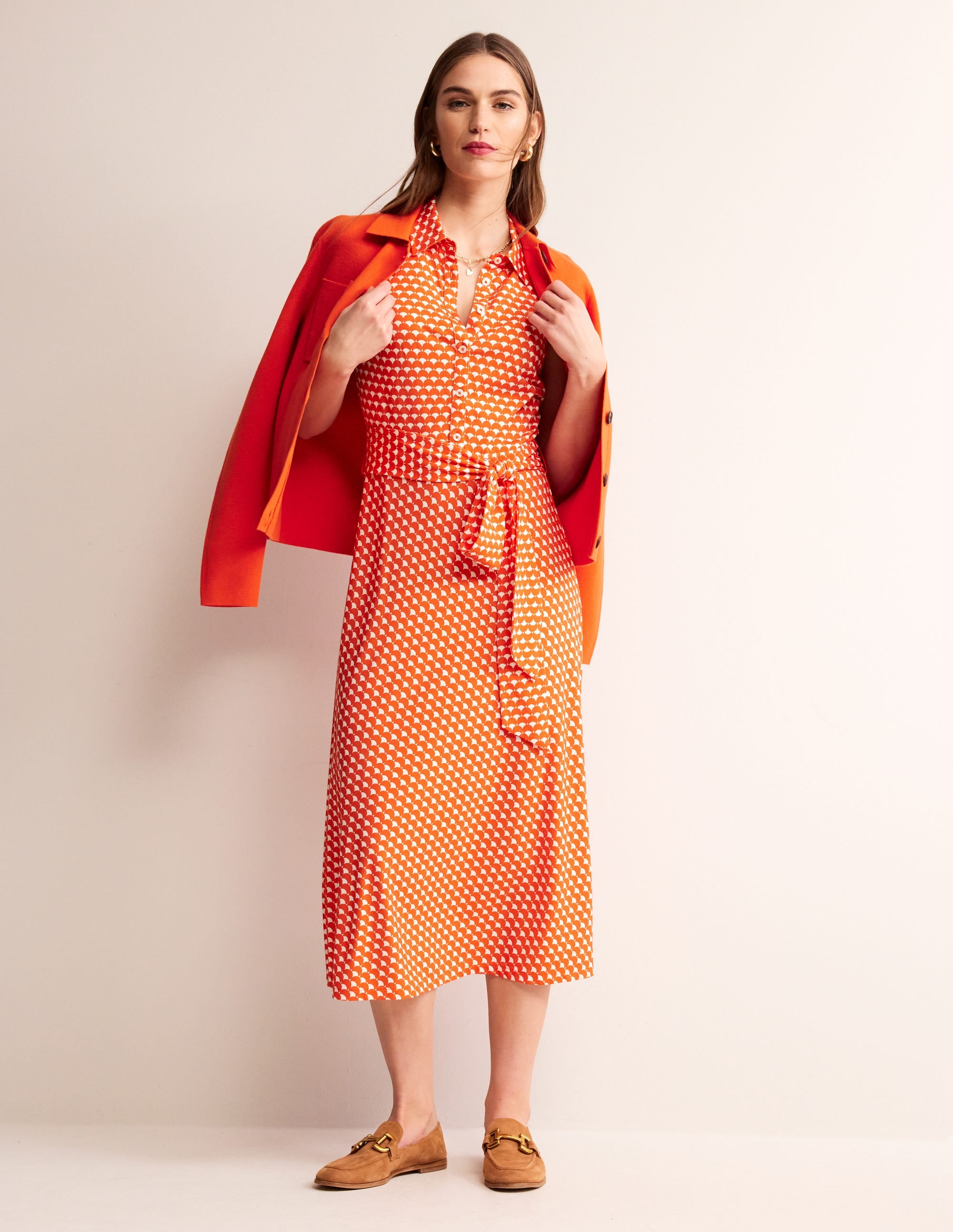 A woman wearing an orange and white crescent stamp sleeveless shirt dress, with a collar and button fastening, paired with a matching orange blazer.