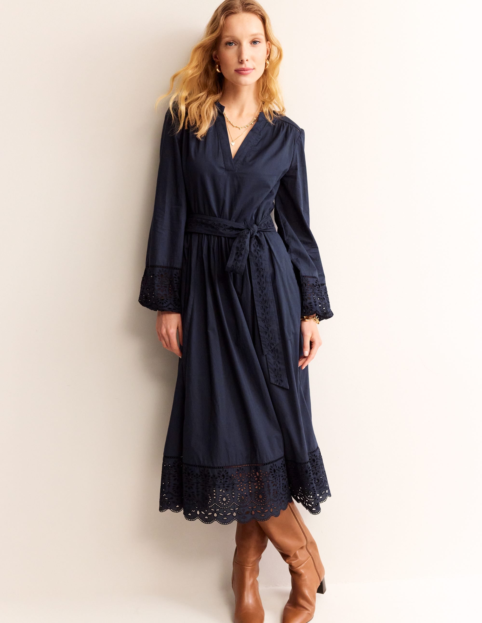 A woman wearing a navy blue midi dress with broderie embroidery, long full sleeves, a belt at the waist, and a scalloped hemline.