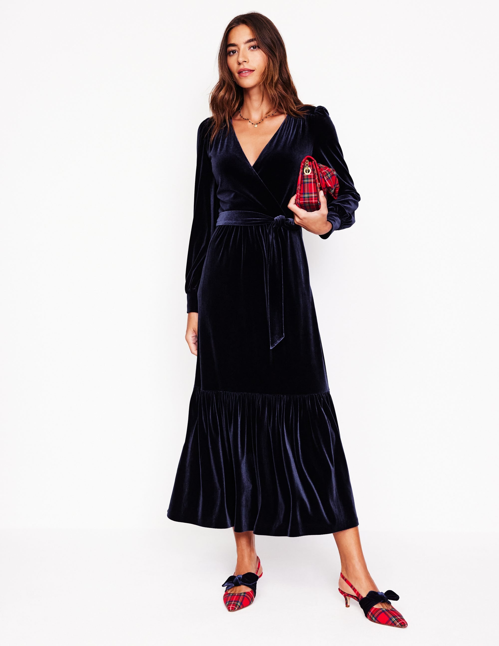 A woman wearing a navy velvet midi wrap dress with a waist belt, pockets, and a flared hem, paired with red plaid heels and holding a red plaid clutch.