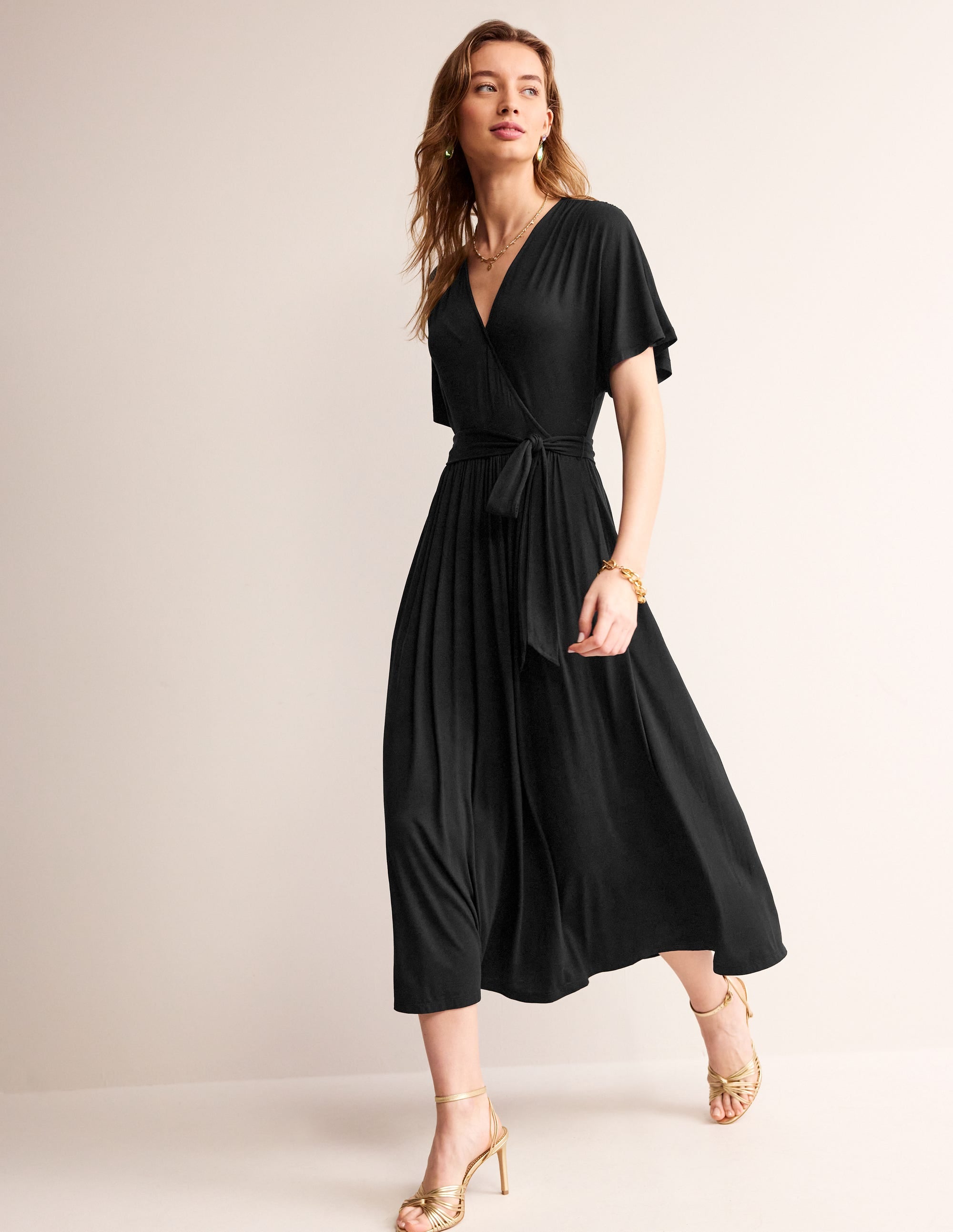 A woman wearing a black kimono wrap midi dress with short sleeves, a waistband, and side pockets, styled with gold heels and jewelry.