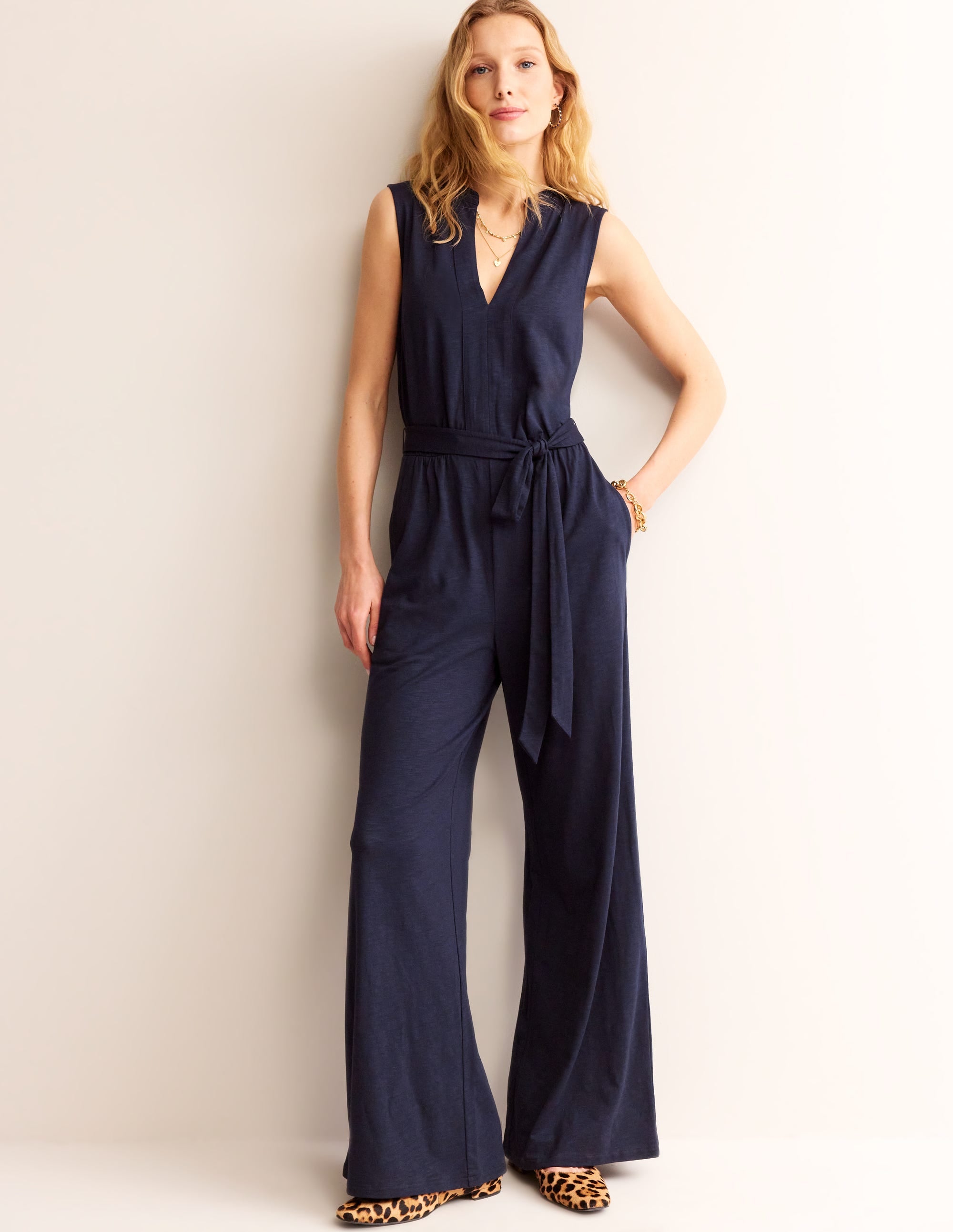 A woman wearing a navy blue jumpsuit with a notch neckline, wide legs, and a self-tie belt, standing against a plain background.