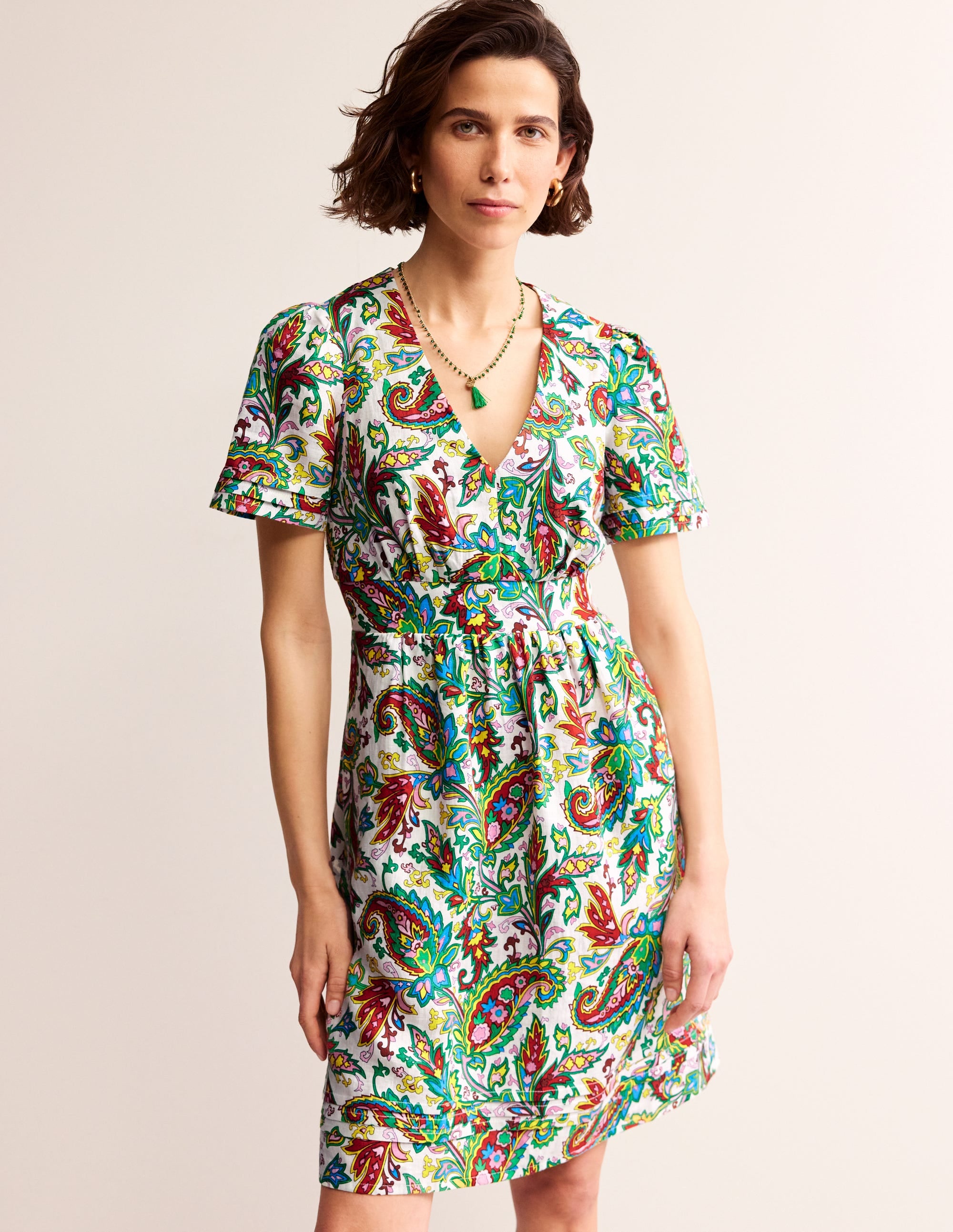 A woman wearing a short, sleeveless linen dress with a vibrant paisley pattern in shades of blue, red, green, and yellow, with puff sleeves and a fitted waist.