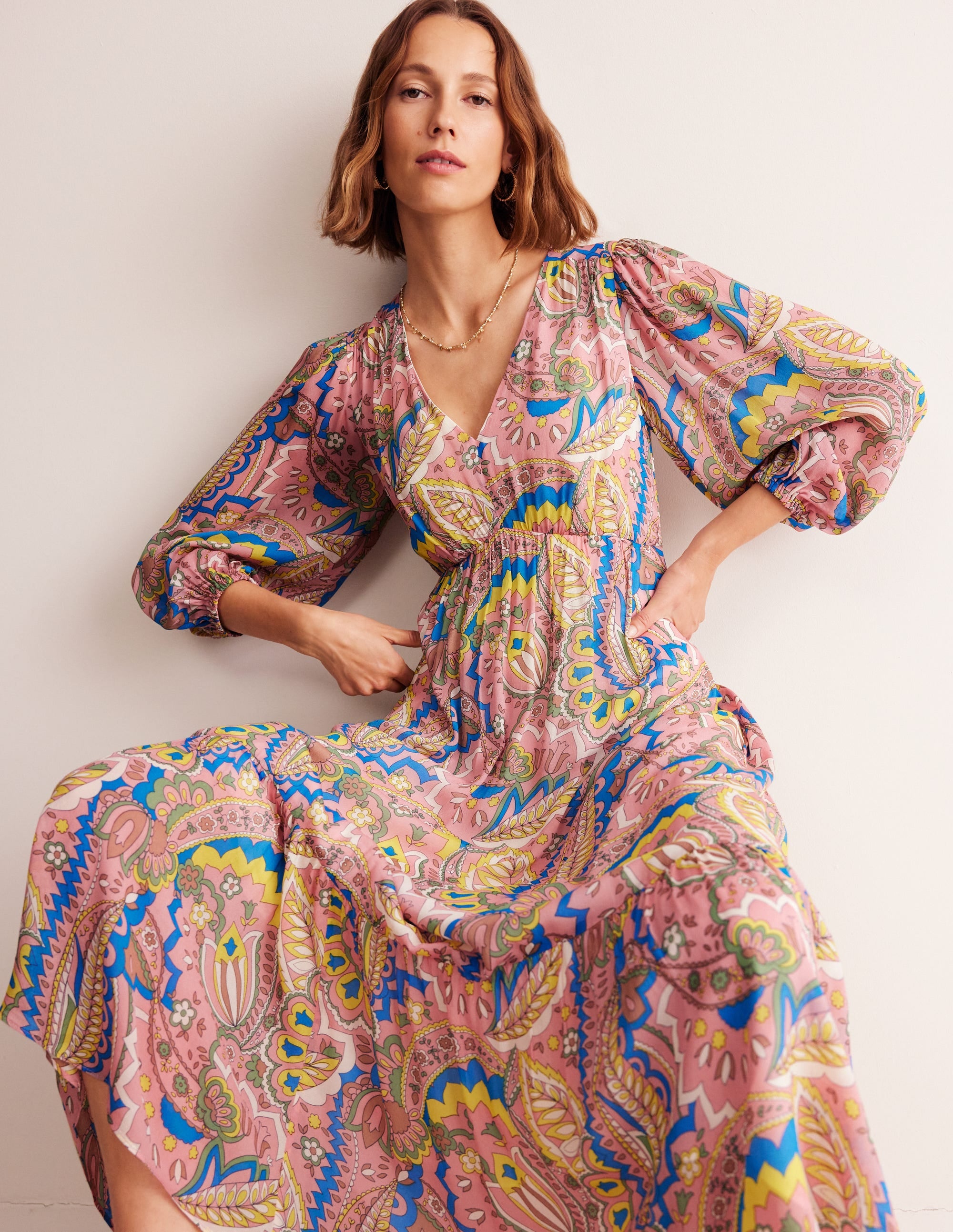 A woman wearing a colorful, patterned maxi dress with a V-neckline, puffed sleeves, and side pockets, standing against a plain background.