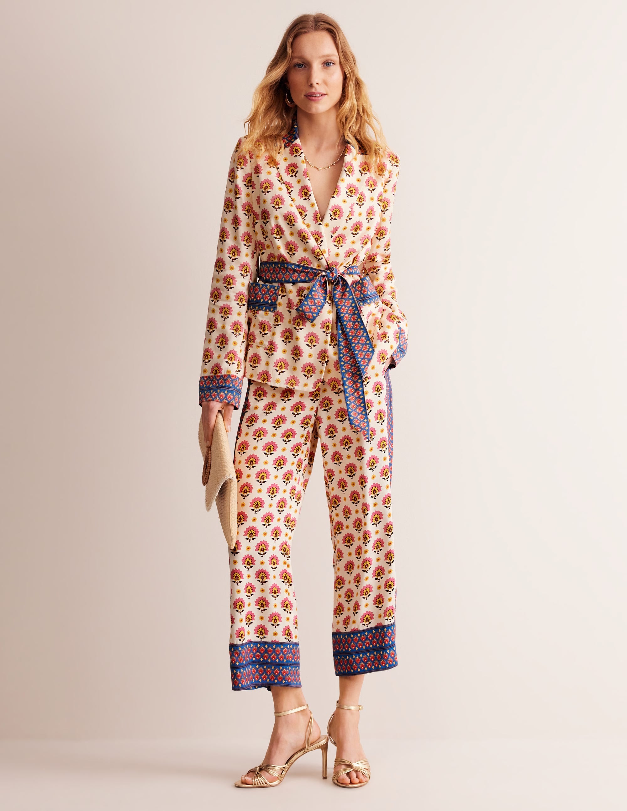 A woman wearing an ivory satin belted blazer with a floral Passion Geo pattern, front patch pockets, and a fitted silhouette.