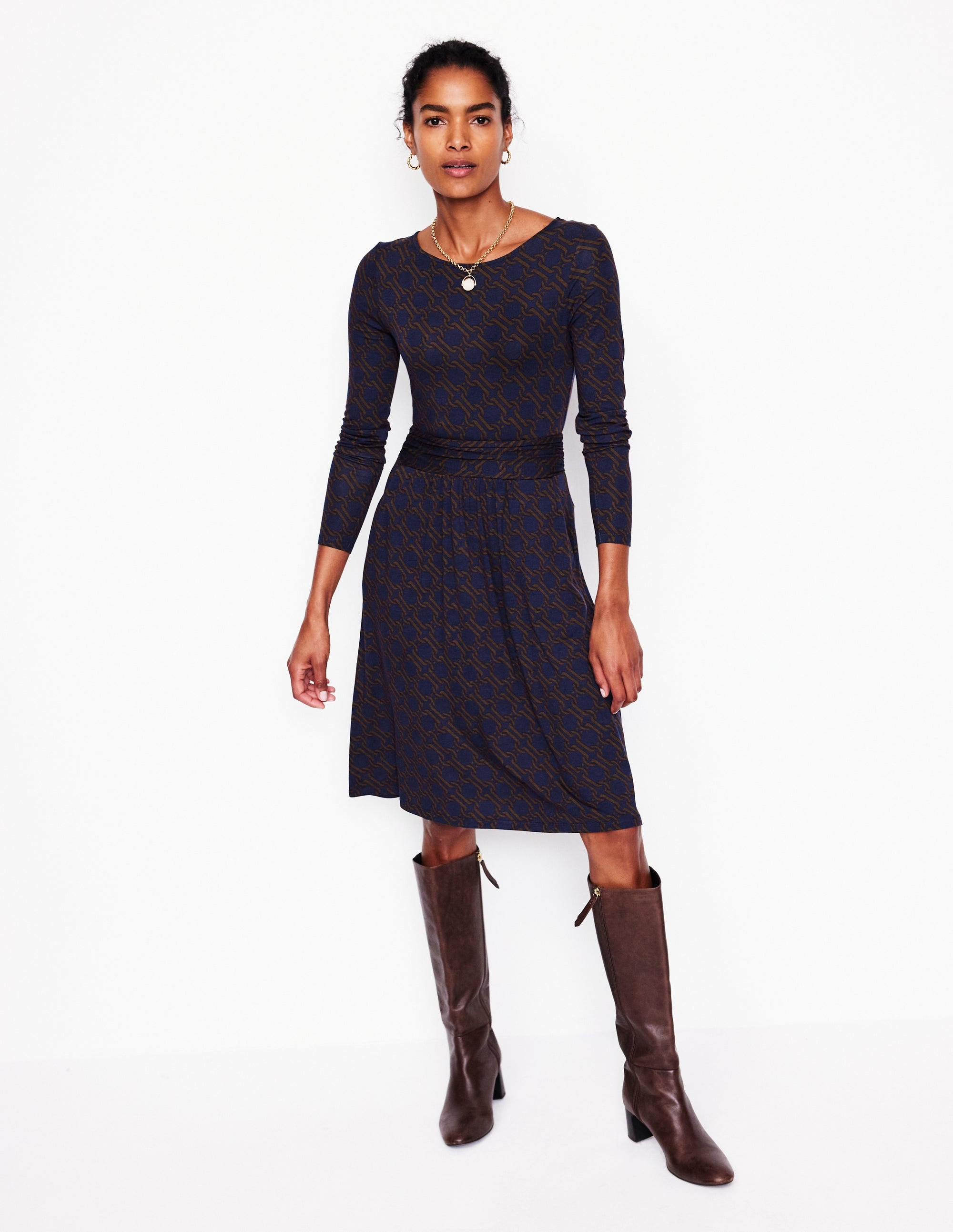 A woman wearing a long sleeve dress with a chain geometric pattern in Raw Umber, featuring a draped fabric and waistline ruching.
