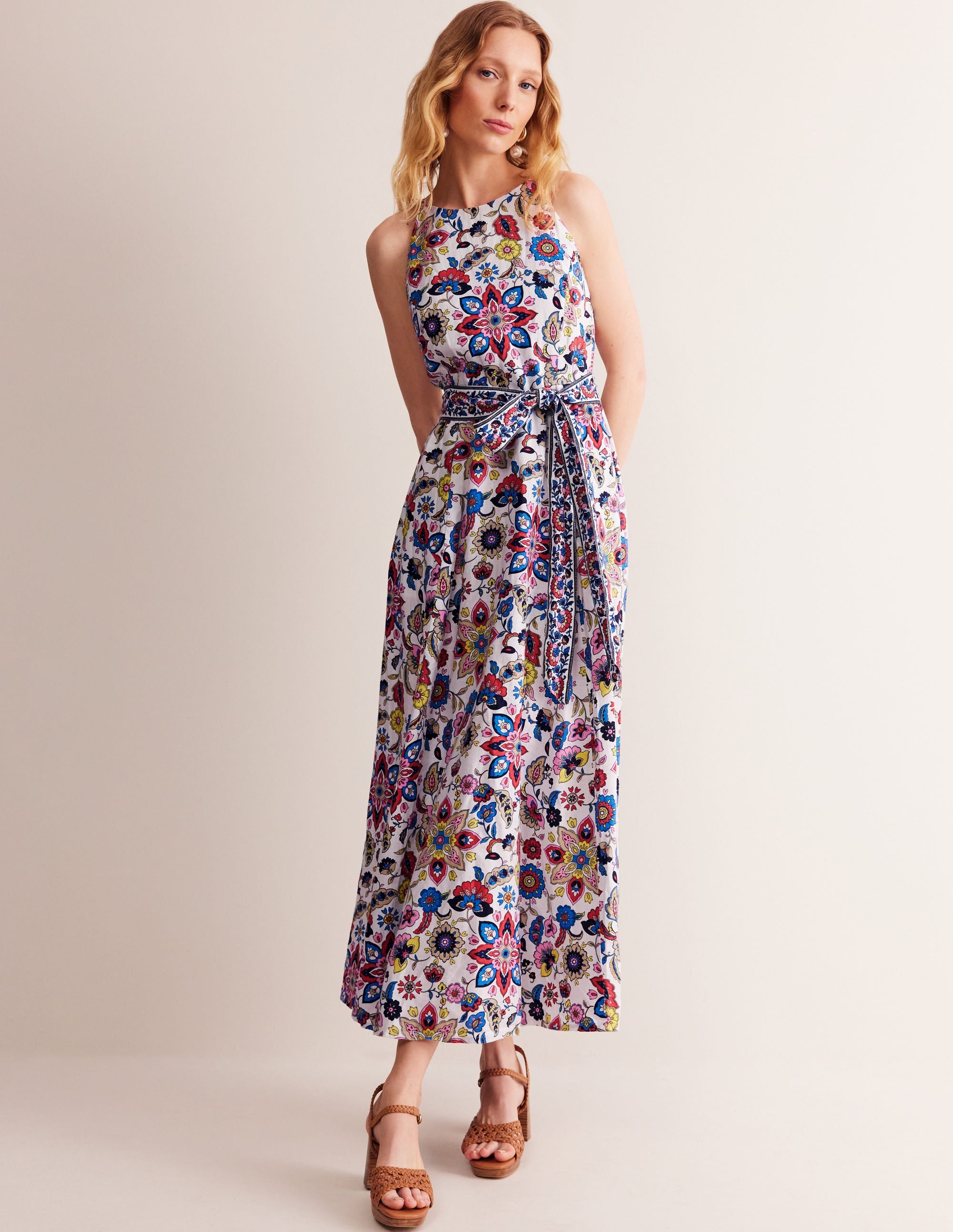 A woman wearing an ivory linen maxi halter dress with a colorful botanical sprig pattern, featuring a longline halterneck, pockets, and a belt.
