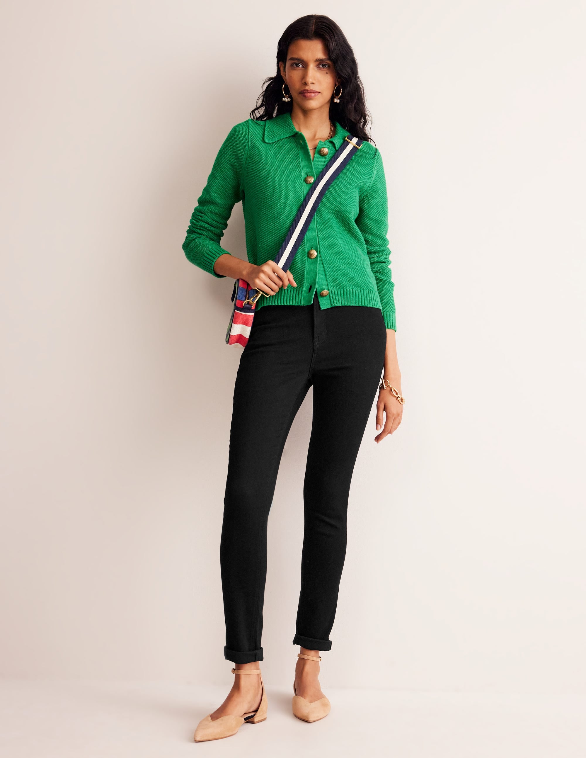 A woman wearing black high-rise skinny jeans, a green sweater, and beige heels, holding a striped bag.