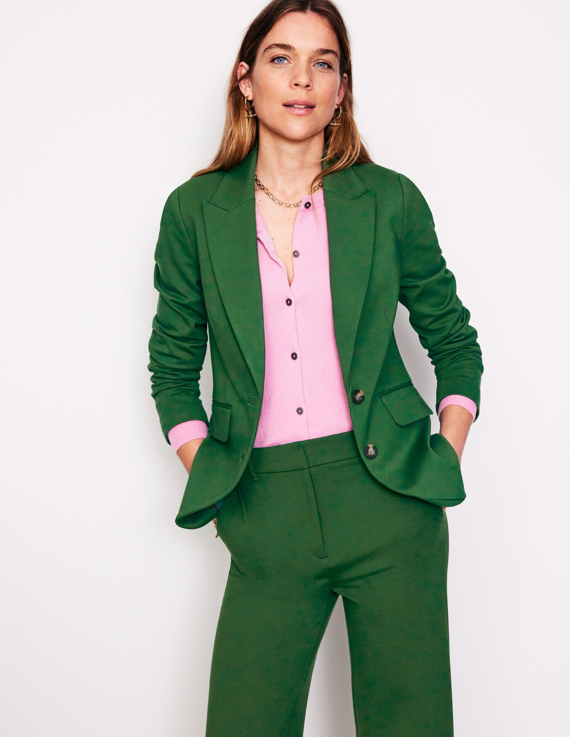 A woman wearing a green blazer with a pink top underneath, standing against a plain background.