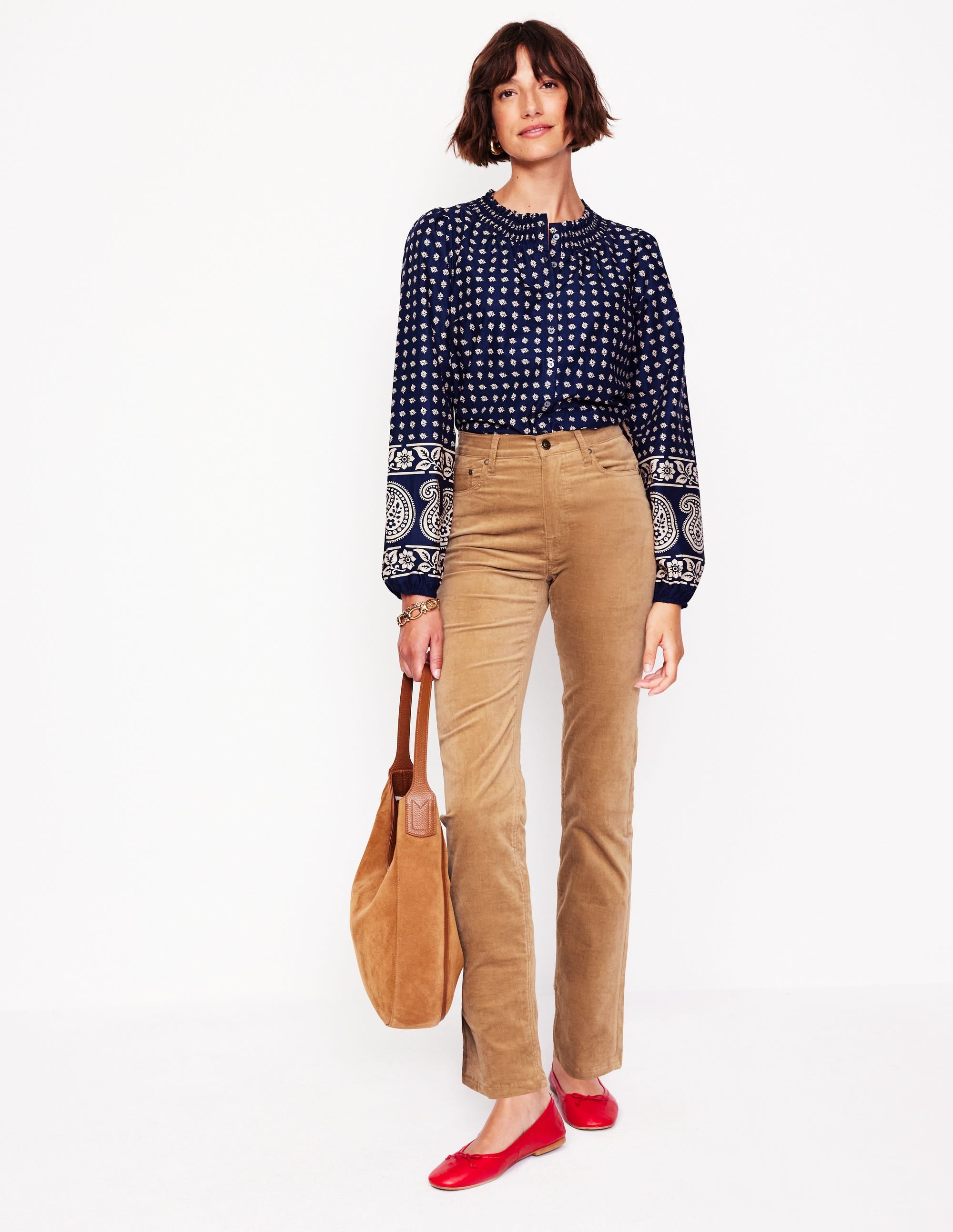 A woman wearing cappuccino-colored velveteen 5 pocket jeans, paired with a navy patterned blouse, red flats, and carrying a tan bag.