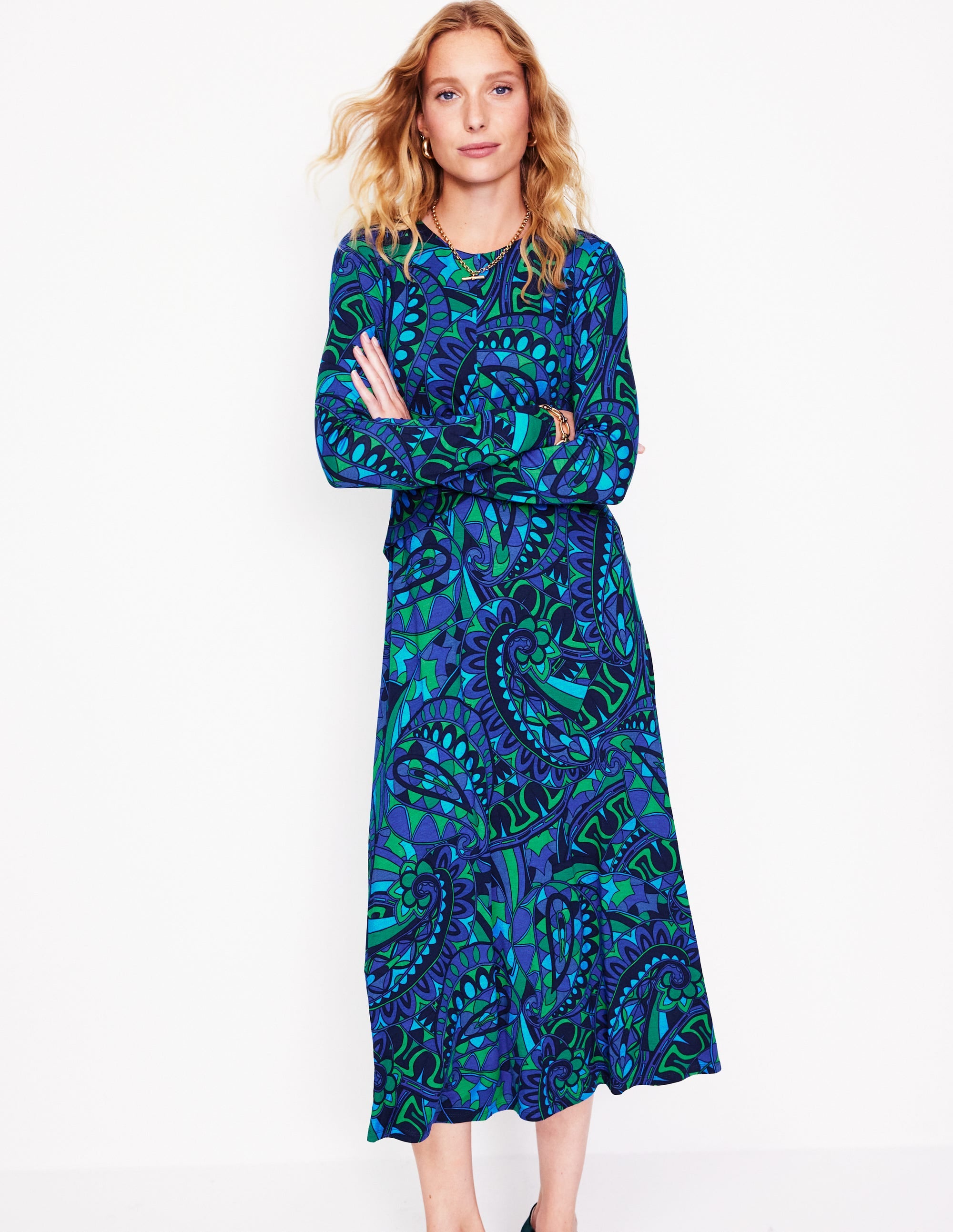 A woman wearing a long, blue and green paisley patterned jersey dress with a twisted waist, lined bodice, and calf-length hem.