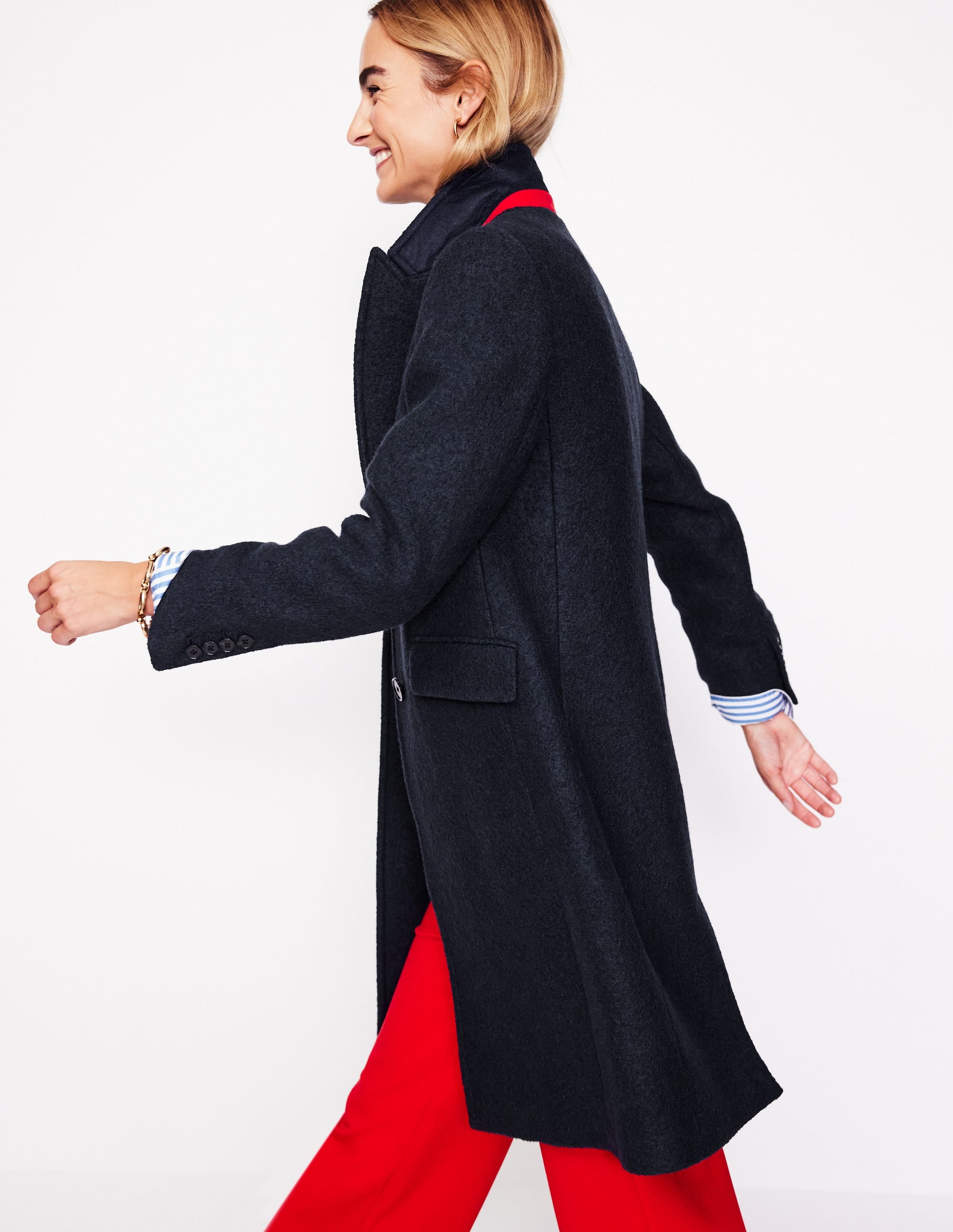 A woman wearing a navy textured coat with lapels, jetted pockets, and a knee-length cut, paired with red pants and a striped shirt.