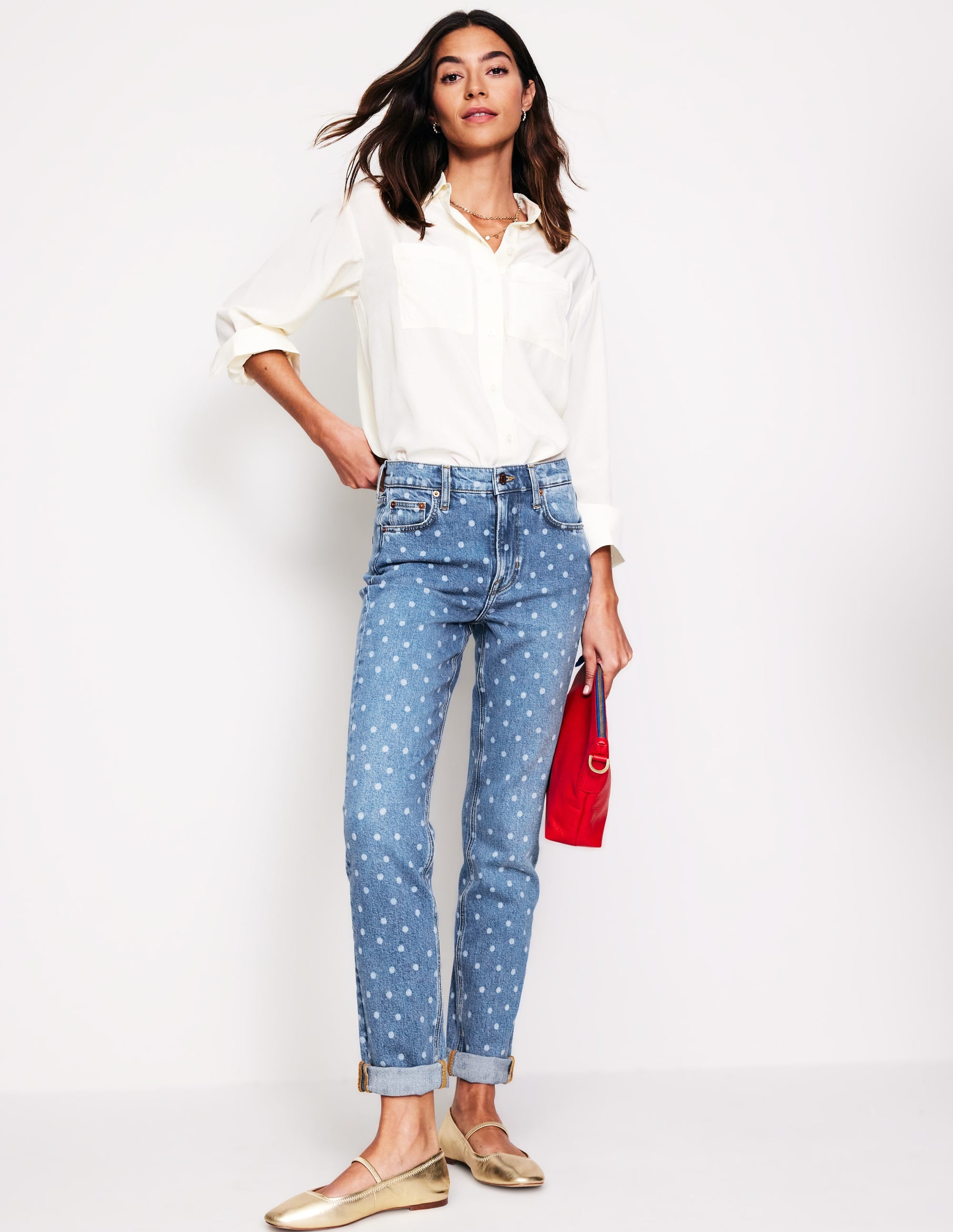 A woman wearing light blue girlfriend jeans with laser spot pattern, a white shirt, and beige flats, holding a red bag.