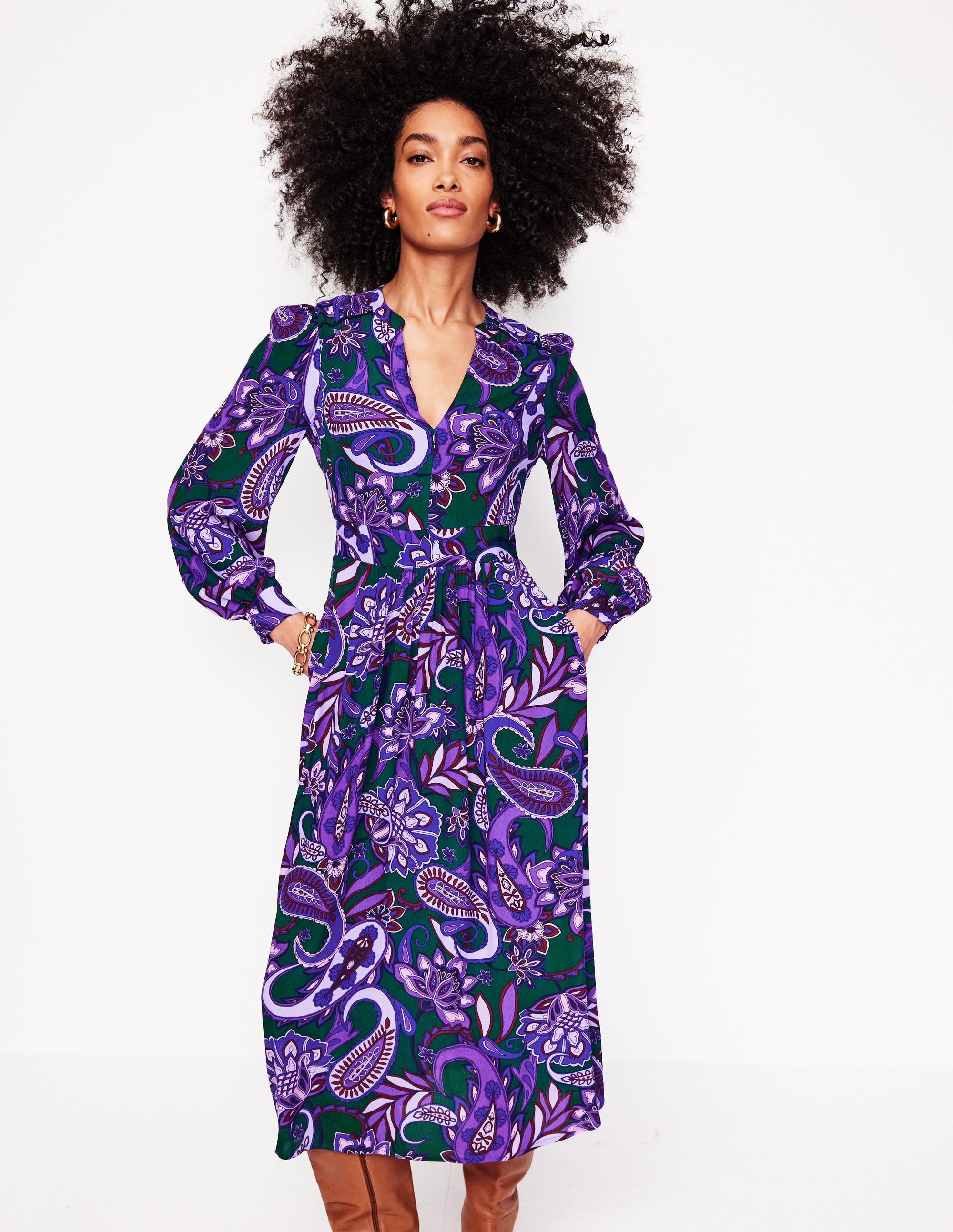 A woman wearing a purple, green, and white paisley patterned midi dress with gathered sleeves and side seam pockets.