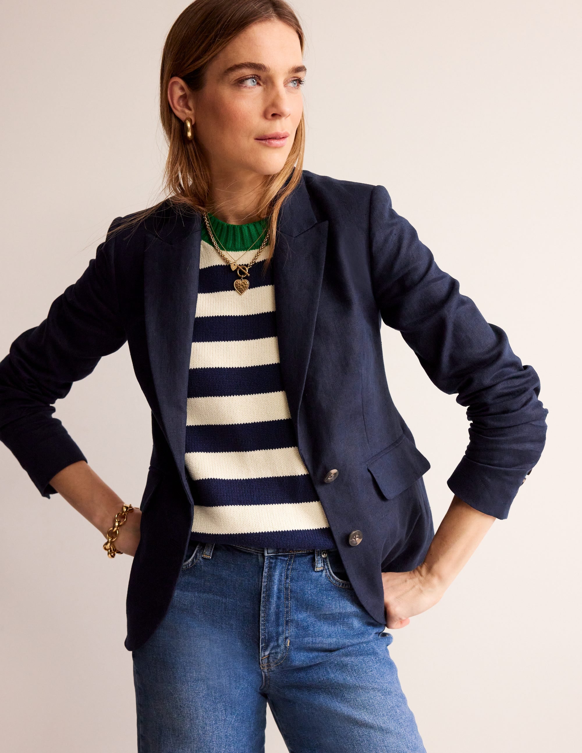 A woman wearing a navy linen blazer with a revere collar, contrast piping, and welt pockets, paired with a striped sweater and jeans.