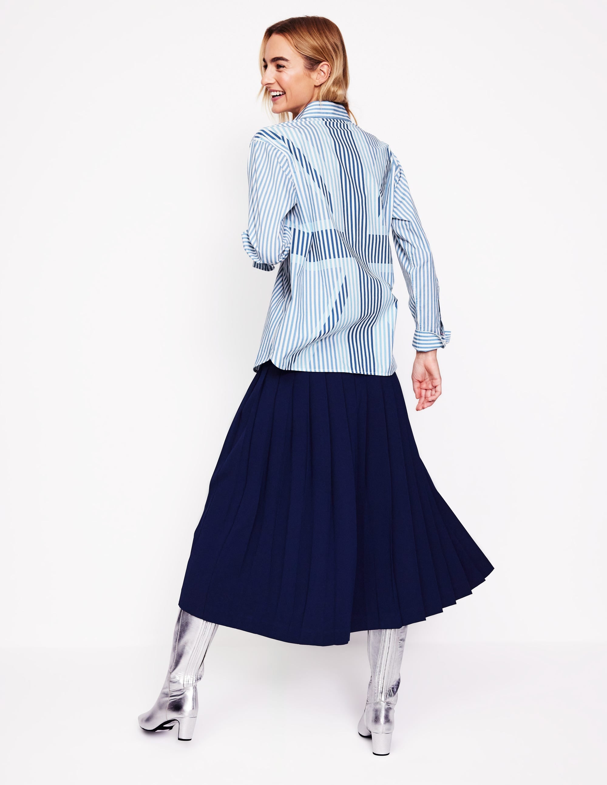 A woman wearing a relaxed-fit cotton shirt with blue and white ticking stripes and a Union Jack pattern, paired with a navy pleated skirt and silver boots.