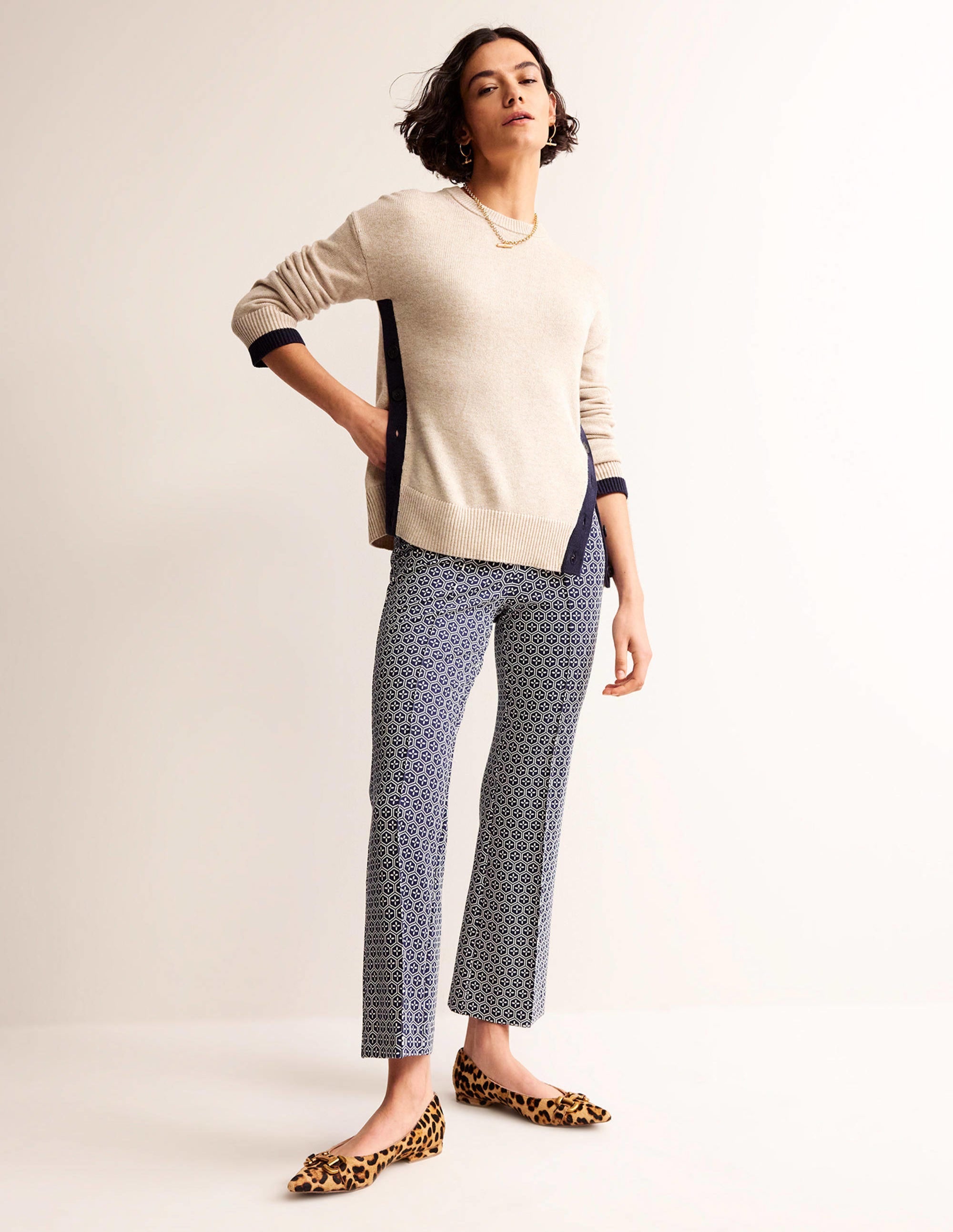 A woman wearing navy blue geo patterned stretch-jersey trousers with a pull-on style and flared leg, paired with a beige sweater and leopard print flats.