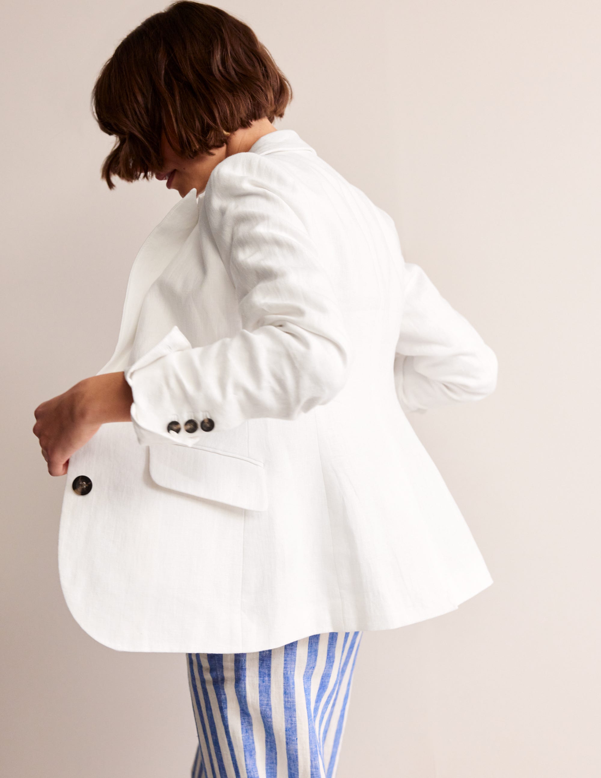 A woman wearing a white linen blazer with a revere collar, contrast piping, and welt pockets, paired with striped pants.