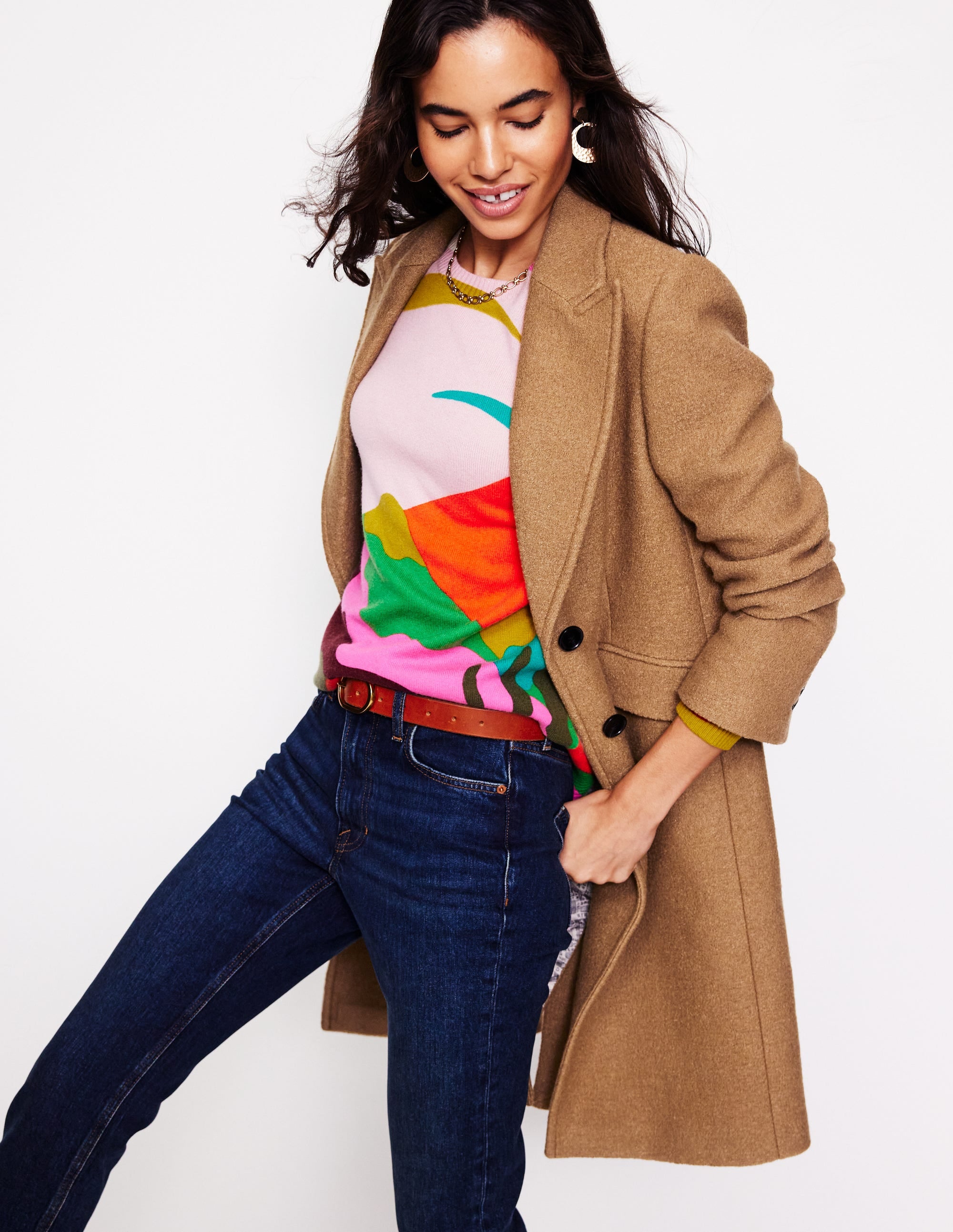 A woman wearing a camel-colored textured coat with lapels and jetted pockets, styled over a colorful abstract print top, with dark blue jeans and accessories.