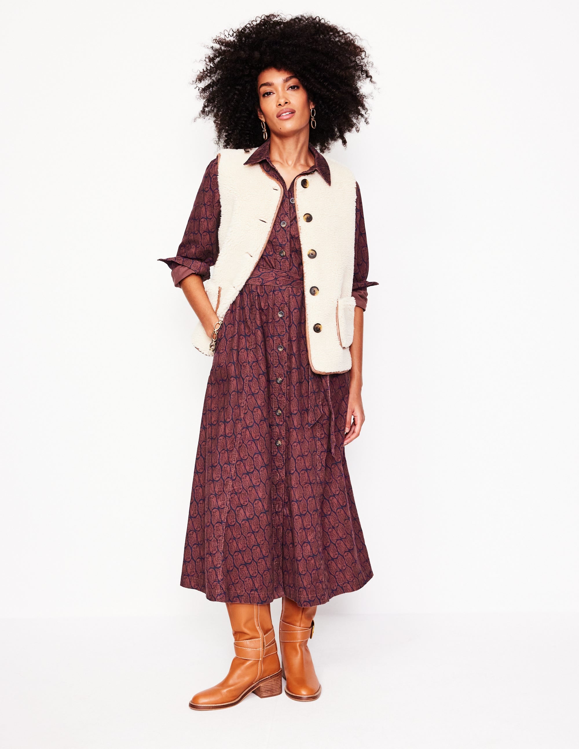A woman wearing a chestnut-colored corduroy midi shirt dress with a button-down front, pleated skirt, and a self-fabric belt, paired with tan boots.