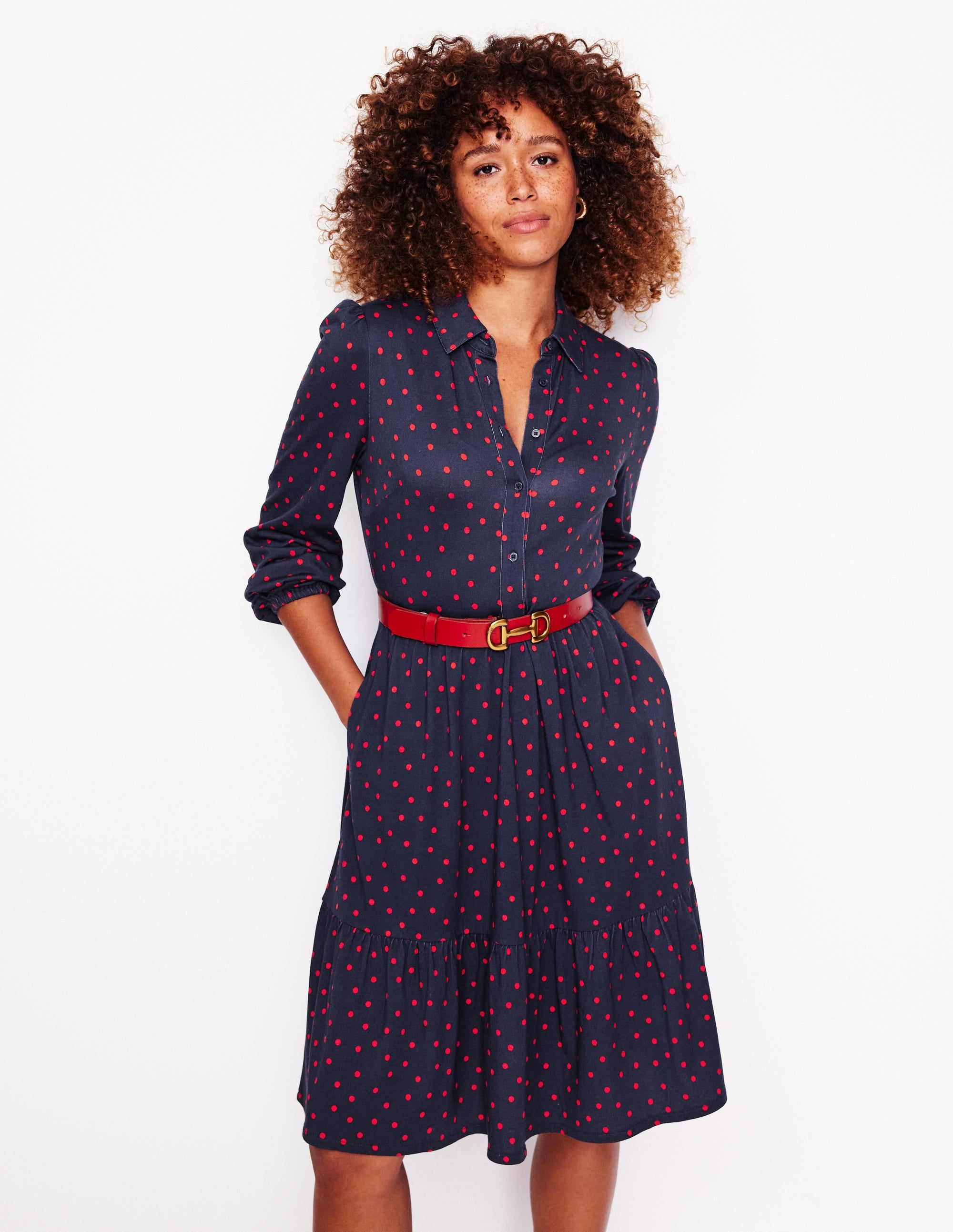 A woman wearing a rouge, tiered jersey dress with sparse spot pattern, featuring a collar, button-down front, and long sleeves rolled up to the elbows.