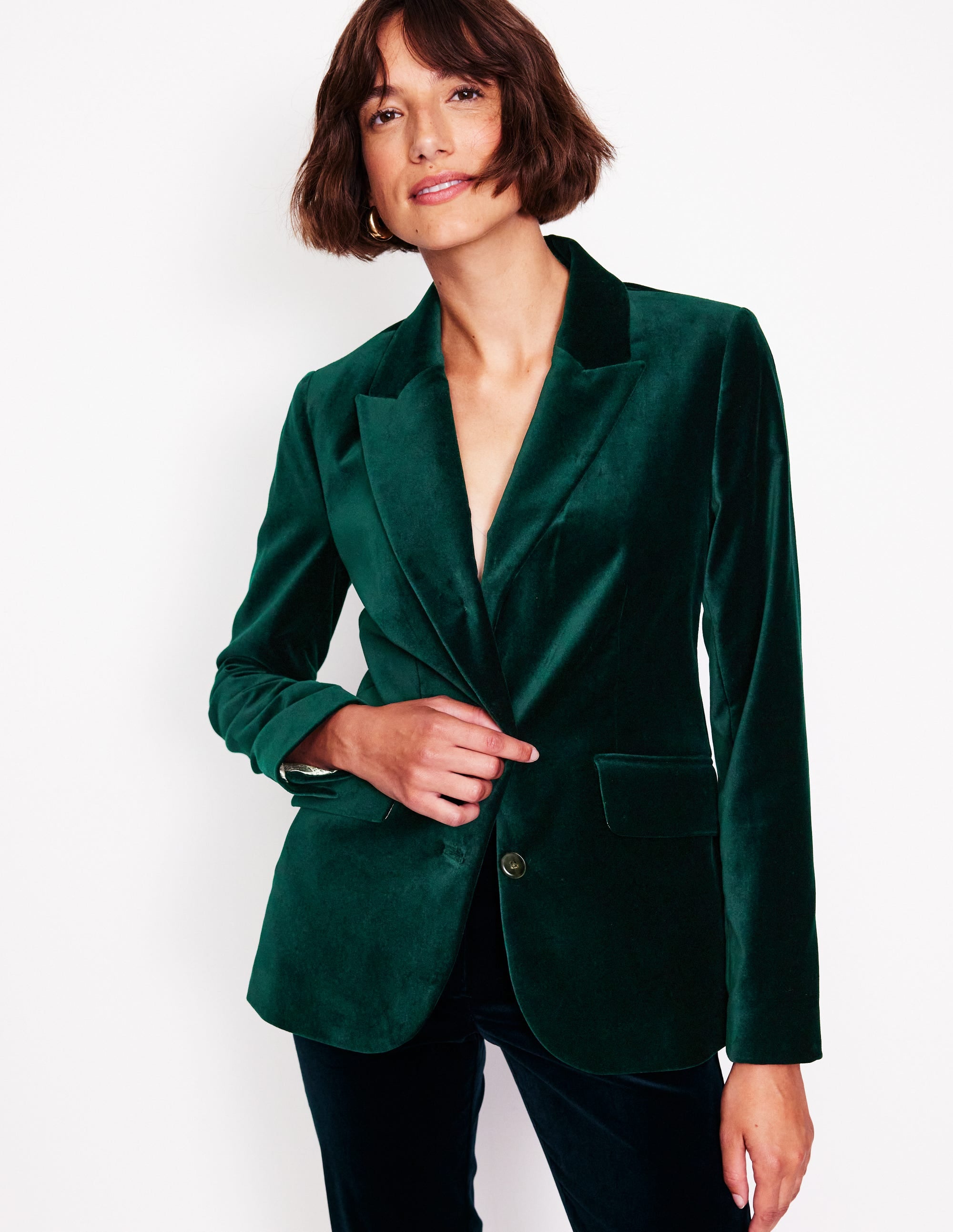 A woman wearing a green velvet blazer with a revere collar, contrast piping, and welt pockets.