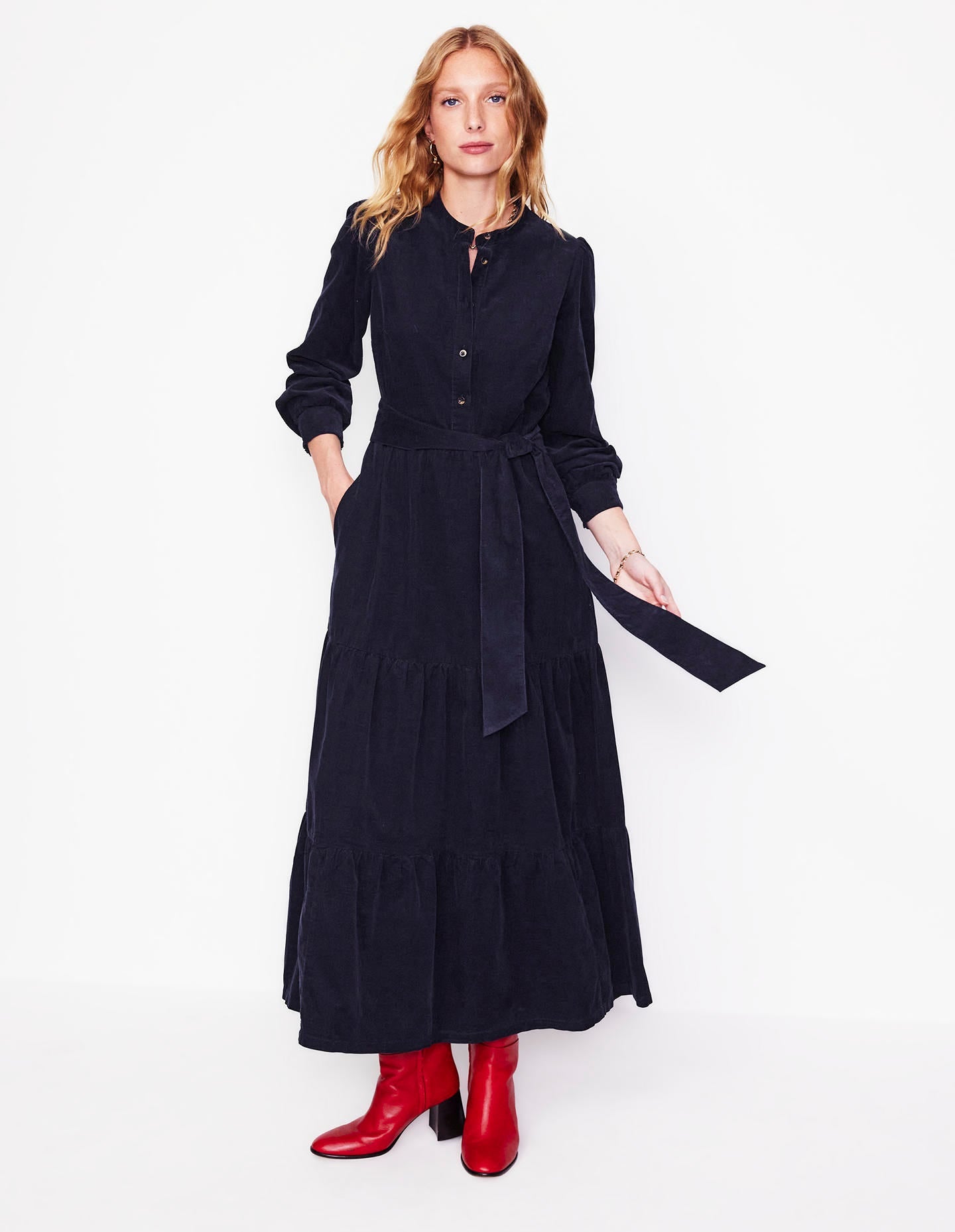 A woman wearing a navy cord maxi dress with long sleeves, tiered design, and a tie waist, paired with red boots.