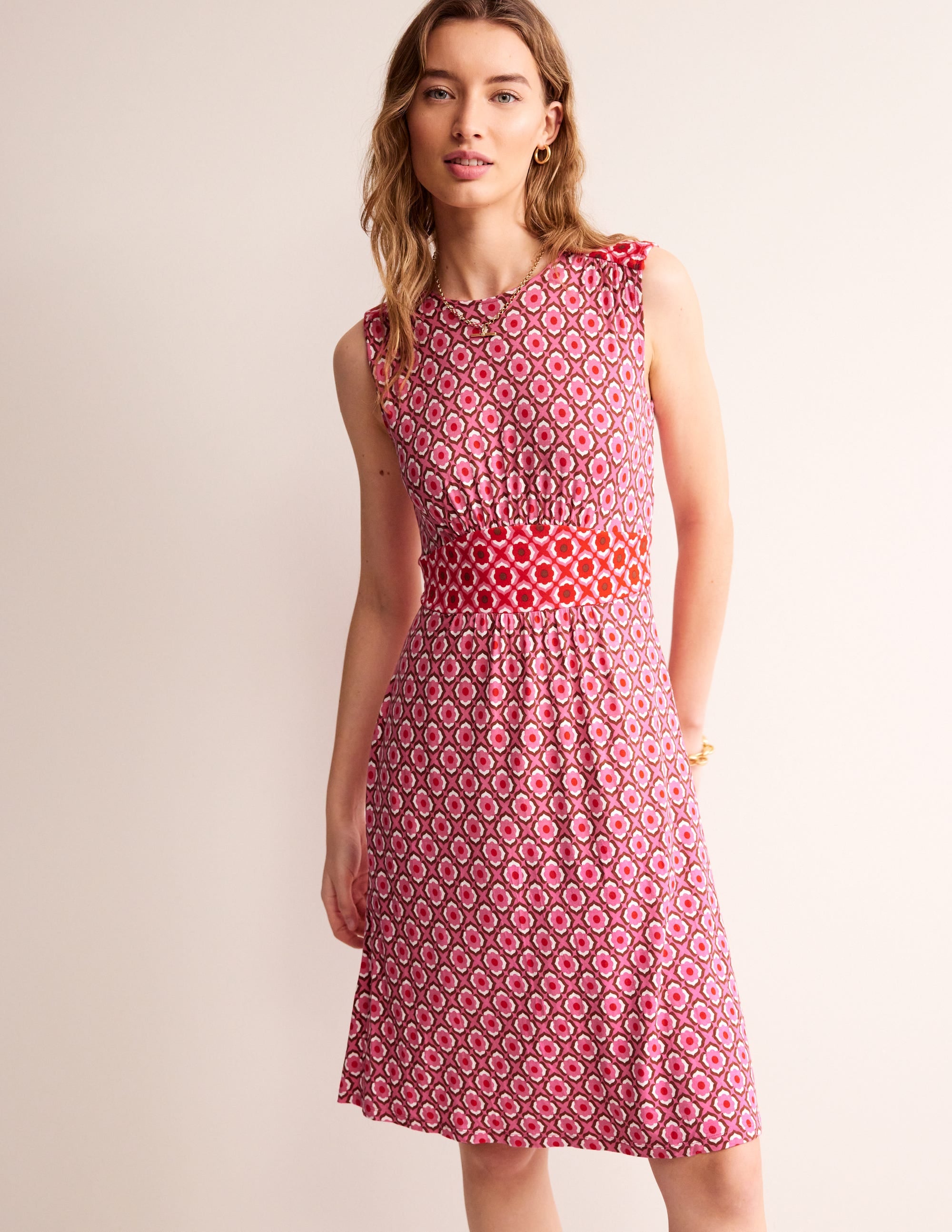 A woman wearing a sleeveless short dress with a colorful geometric pattern, flowing fabric, and knee-length cut.
