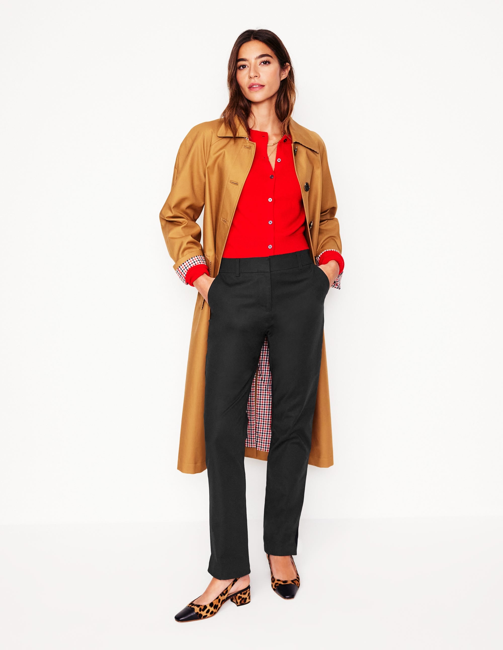 A woman wearing black tapered trousers with a fitted waist, relaxed fit over hips and thighs, and ankle length, paired with a red top and a tan trench coat.