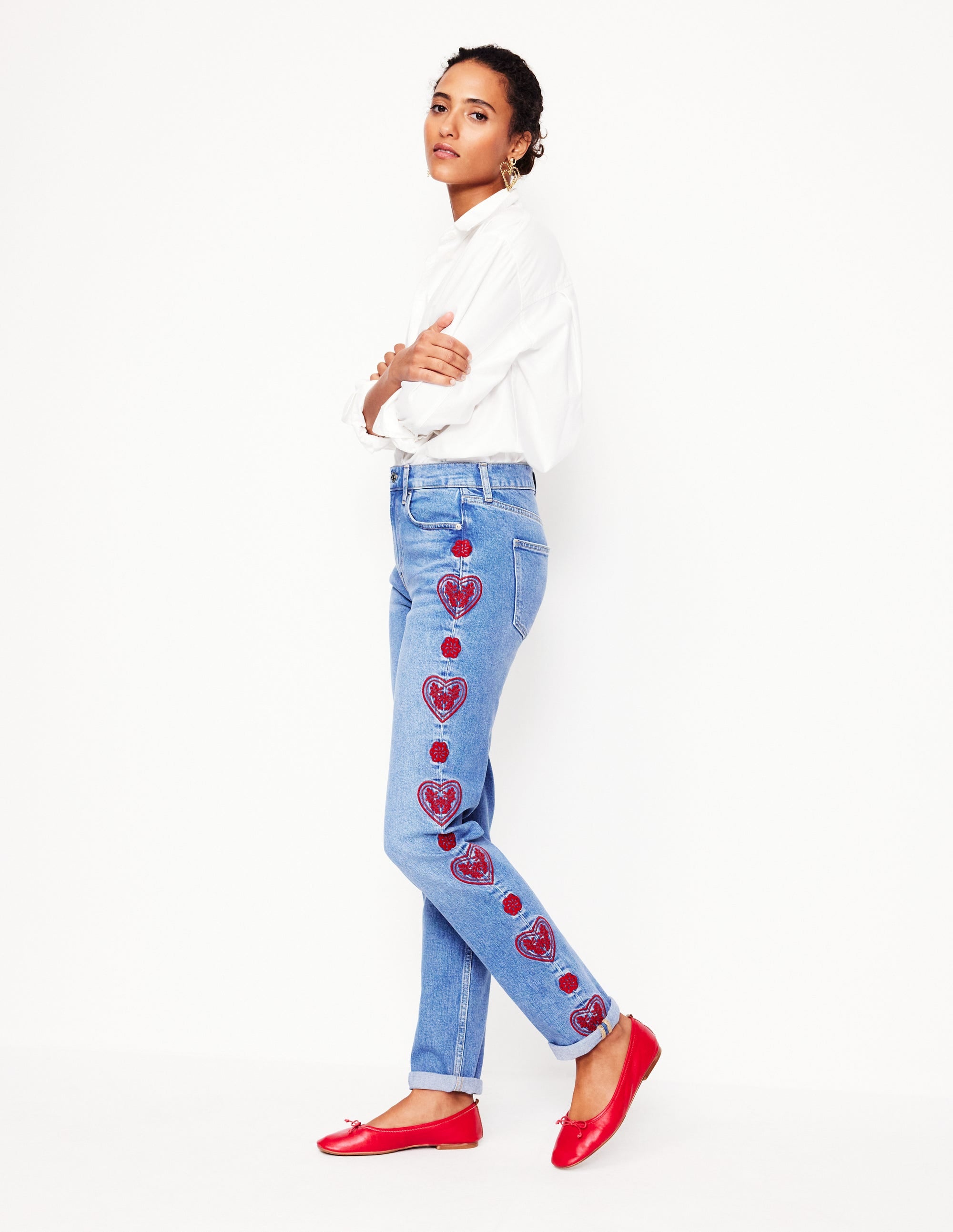 A woman wearing light blue girlfriend jeans with red heart embroidery on the side, paired with a white shirt and red flats.