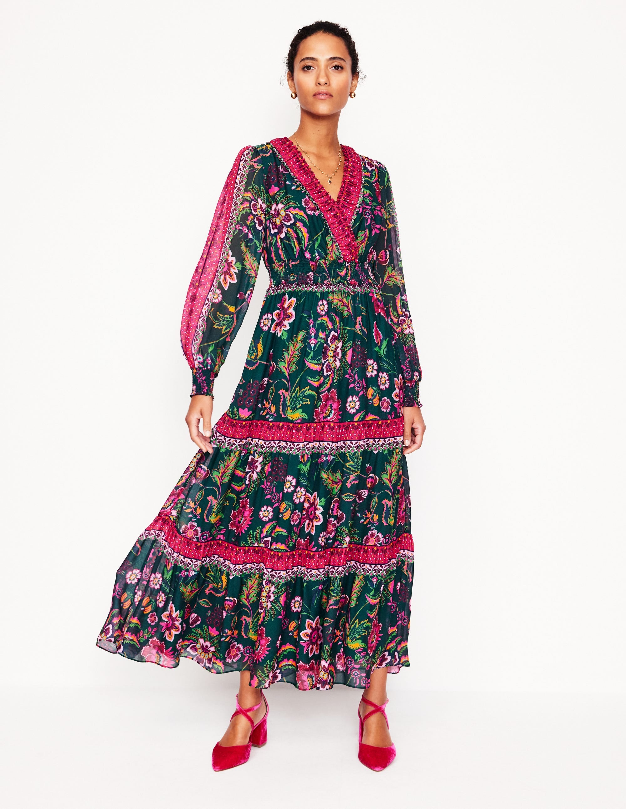 A woman wearing a multicolour floral maxi dress with a V-neckline, long semi-sheer sleeves, and a tiered skirt, standing against a plain background.