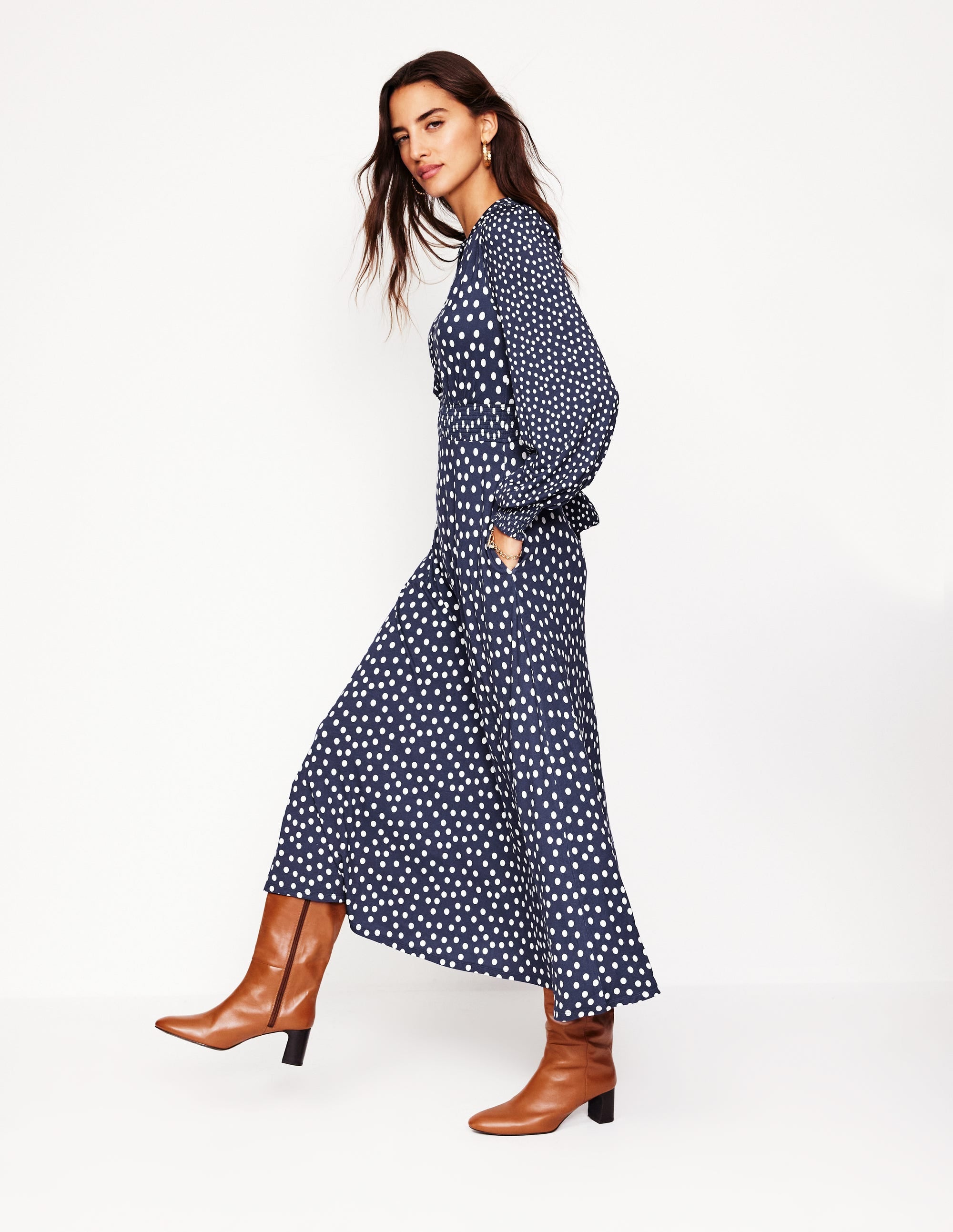 A woman wearing a navy blue midi dress with white abstract dots, featuring a notch neckline and smocked waist, paired with brown knee-high boots.