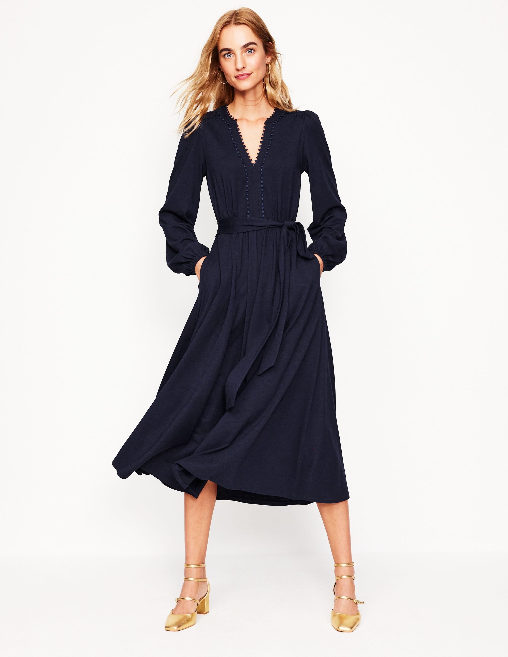 A woman wearing a navy midi dress with long sleeves, a V-neckline with decorative trims, and a self-fabric belt, standing against a plain background.