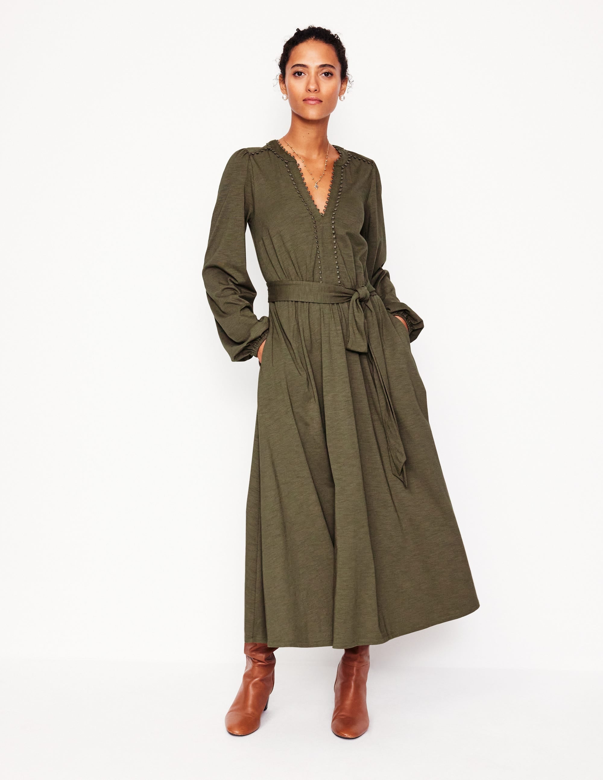 A woman wearing a khaki midi dress with subtle trims, long sleeves, a V-neckline, side pockets, and a fabric belt, paired with brown boots.