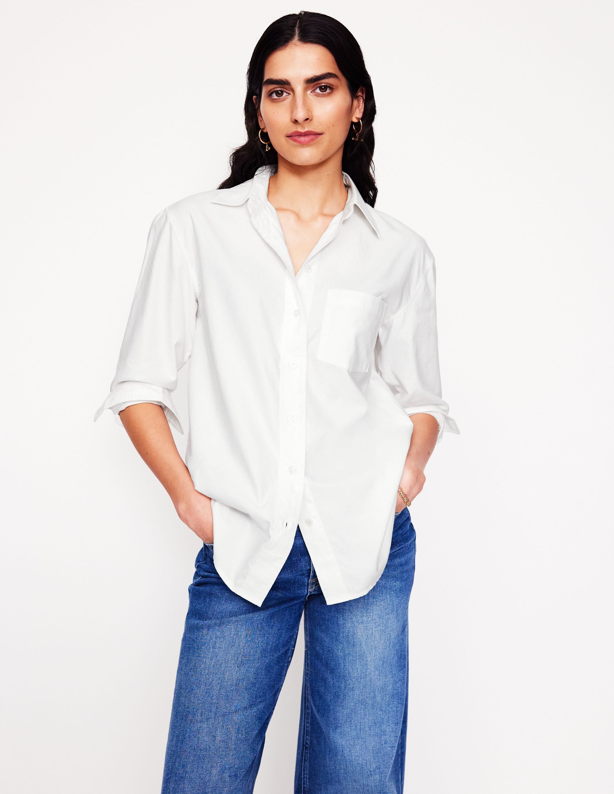 A woman wearing a white oversized cotton shirt with a collar, long sleeves rolled up, and a front pocket, paired with blue jeans.