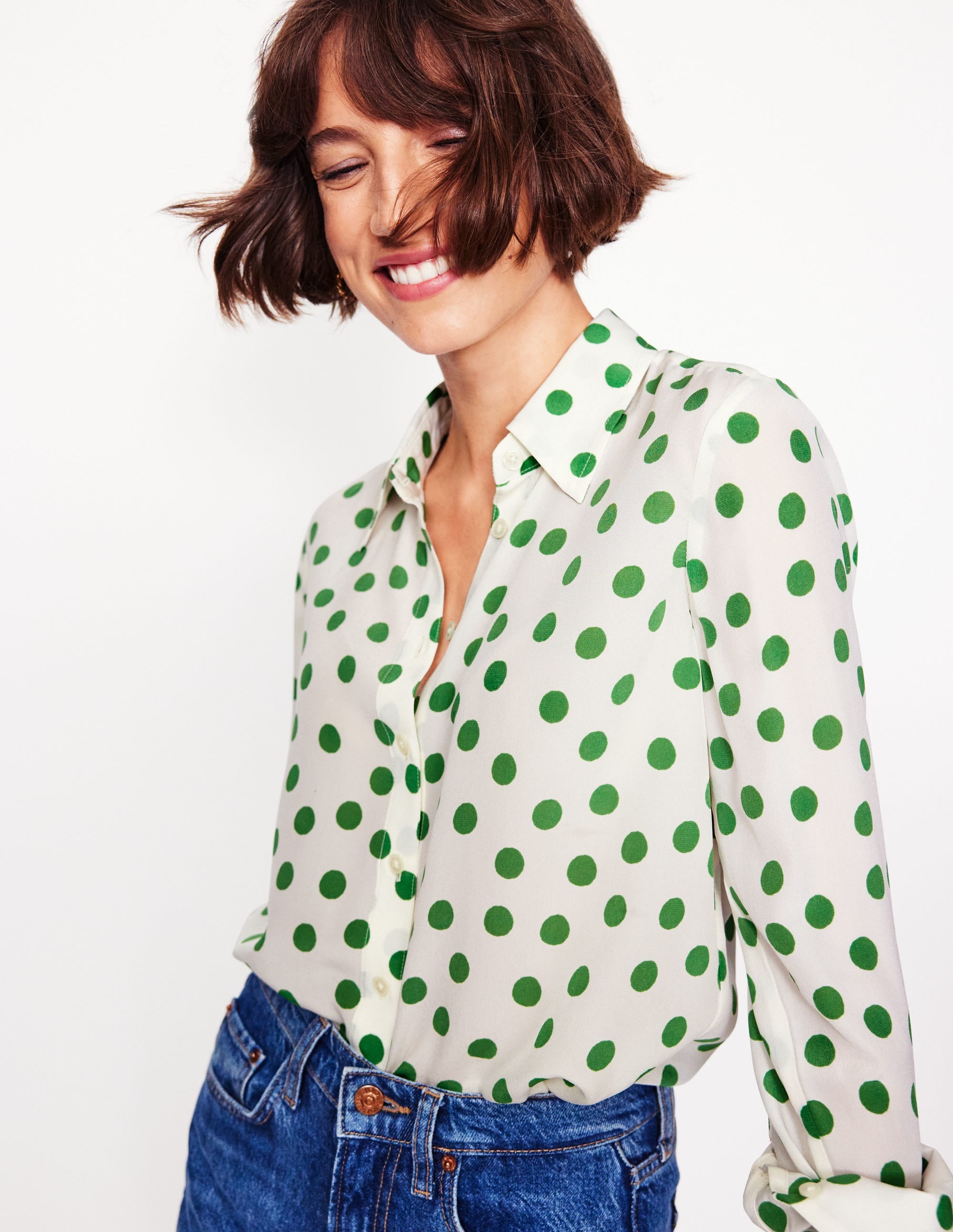 A woman wearing a white silk shirt with green abstract dots, with a collar and button-down front, paired with blue jeans.