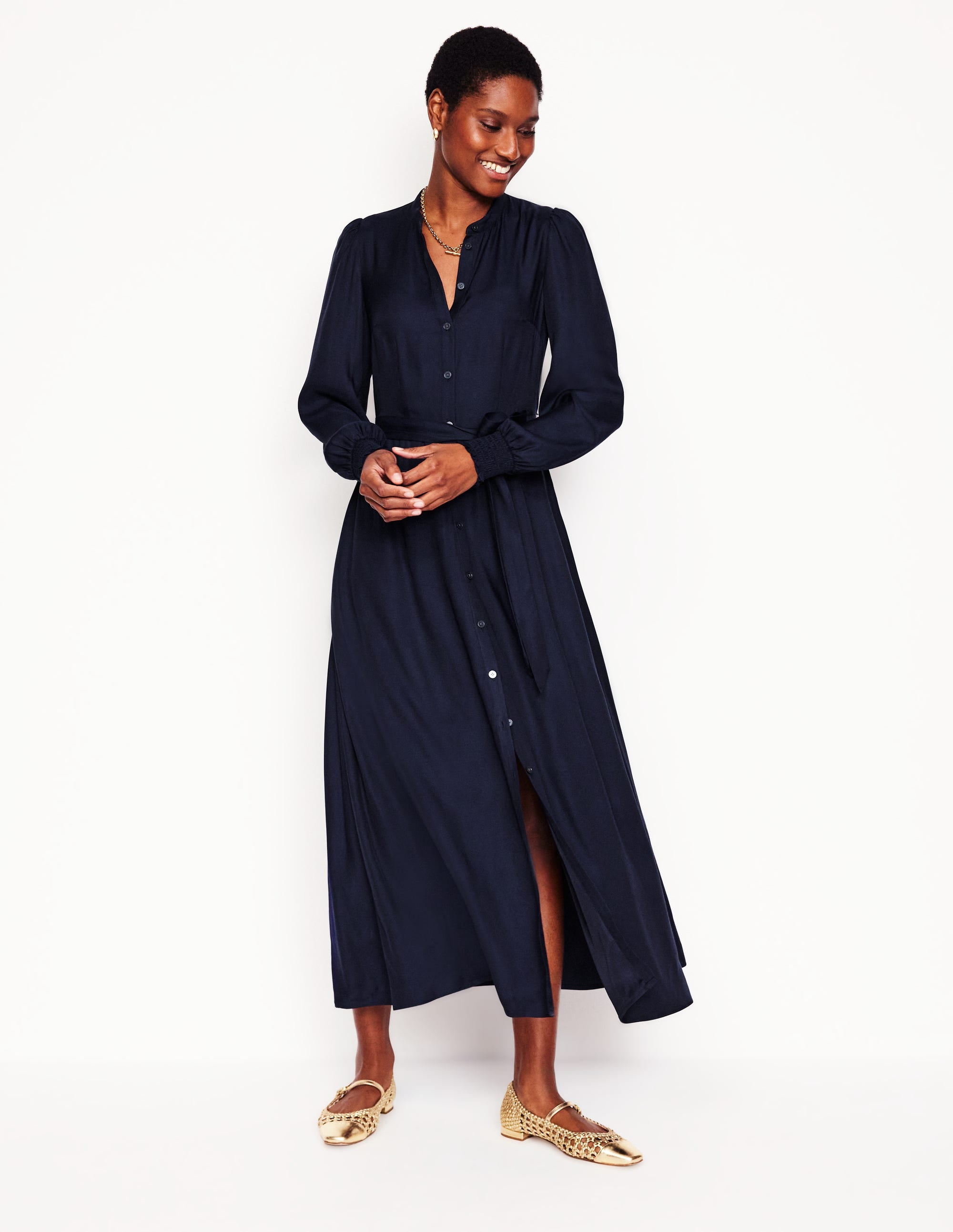 A woman wearing a navy collarless shirt dress with long sleeves, a tie waist belt, and a full skirt with pockets. She is smiling and standing against a plain background.