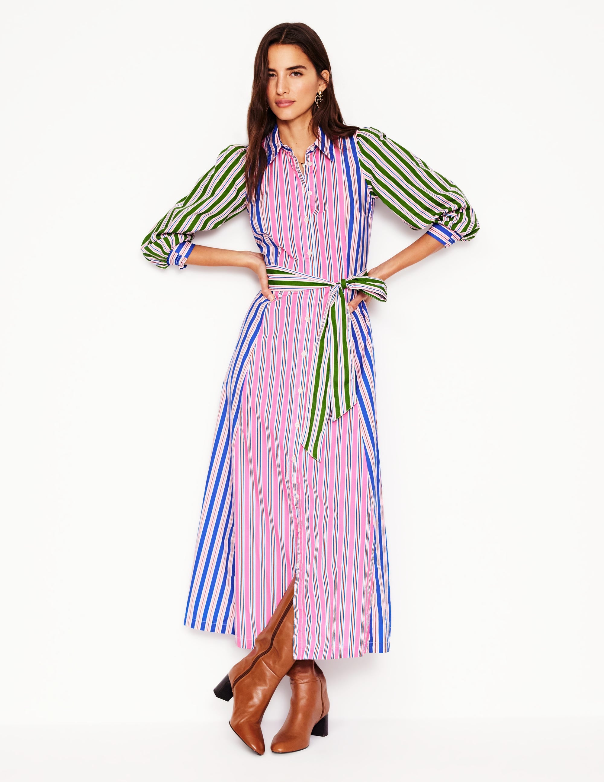 A woman wearing a long, colorful striped shirt dress with a belt, pockets, and brown boots.