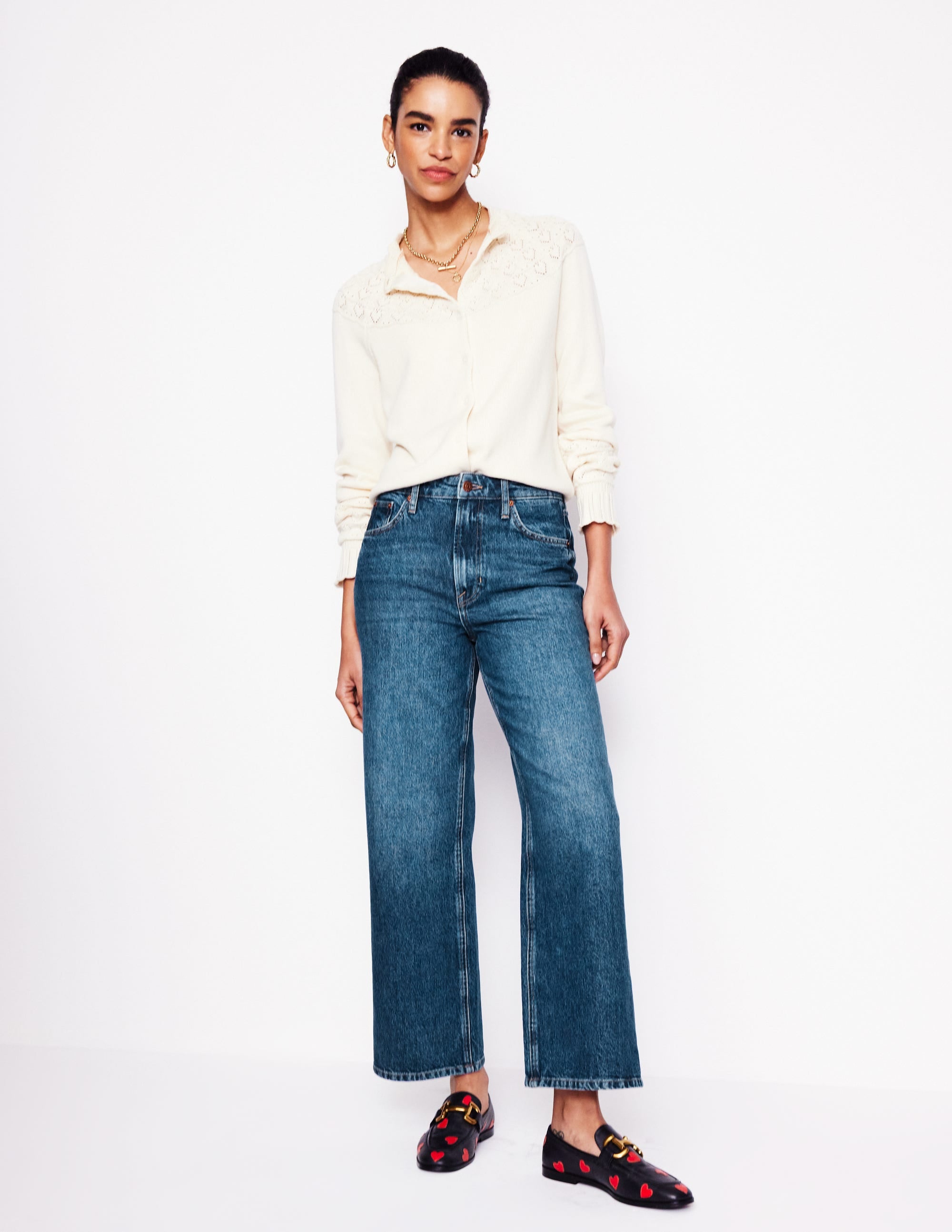 A woman wearing high-rise straight crop jeans in mid vintage blue denim, paired with a cream blouse and black loafers with red heart patterns.
