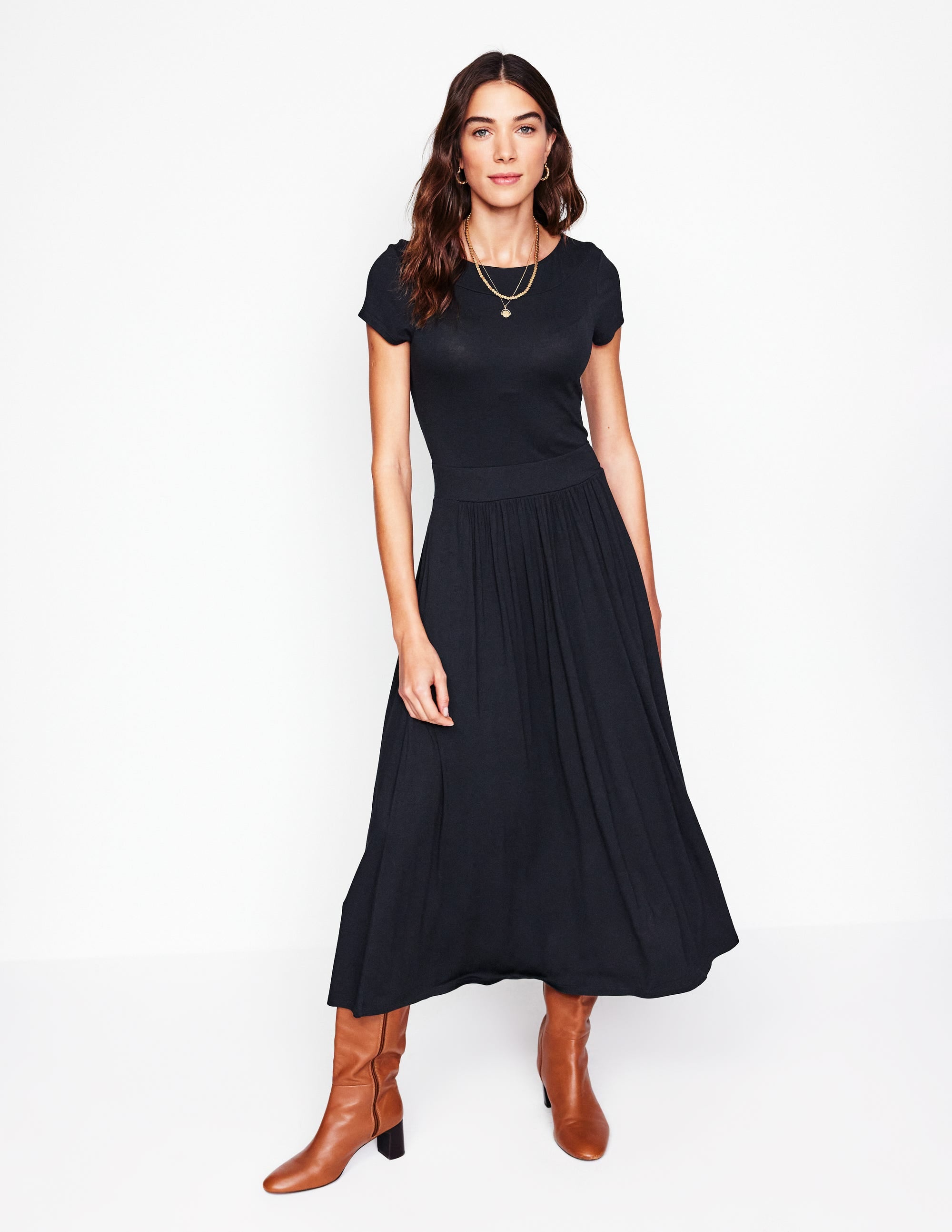 A woman wearing a black midi dress with short sleeves, a wide waistband, and side pockets, paired with brown knee-high boots.