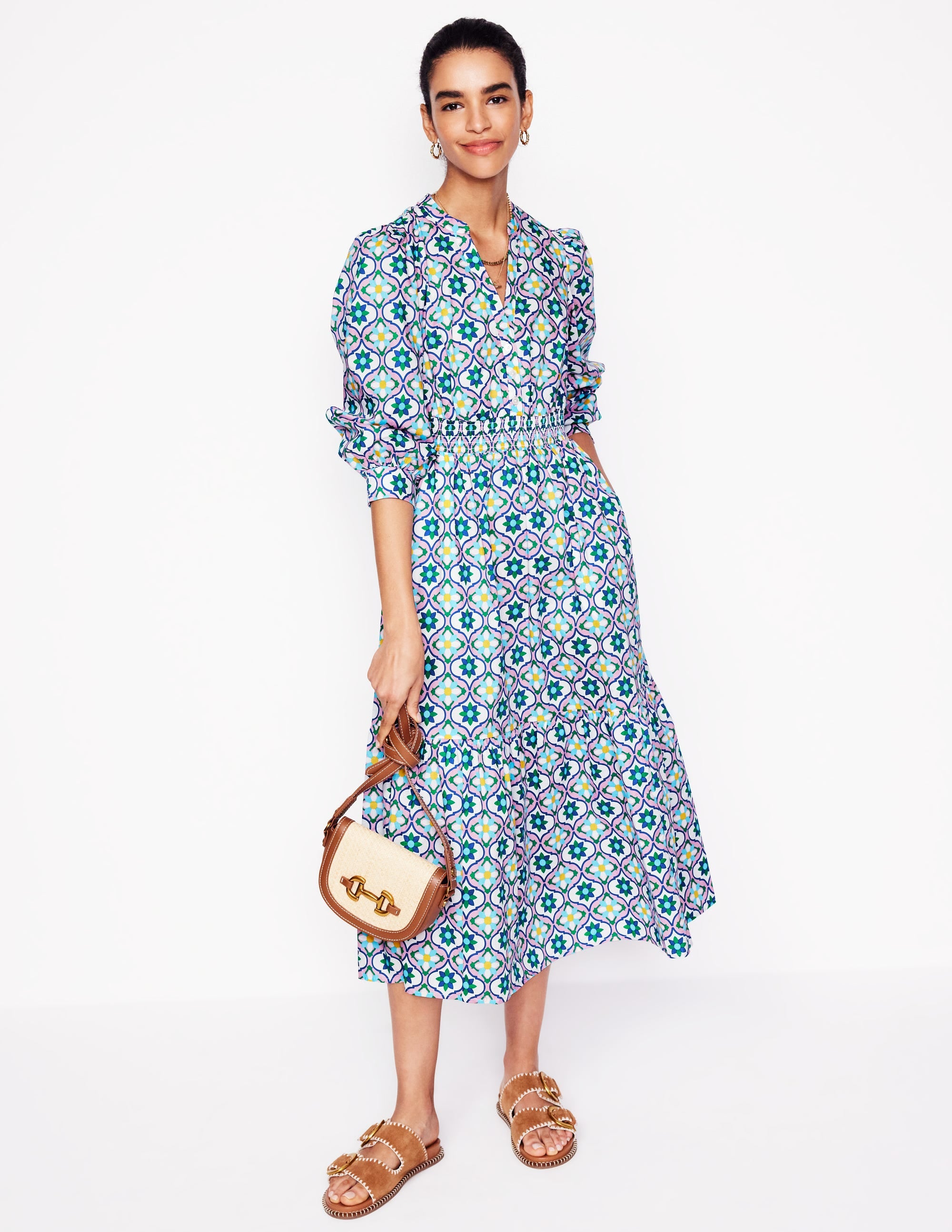 A woman wearing a colorful floral tile patterned midi dress with long puffed sleeves, V-neckline, and side pockets, holding a small handbag and wearing sandals.