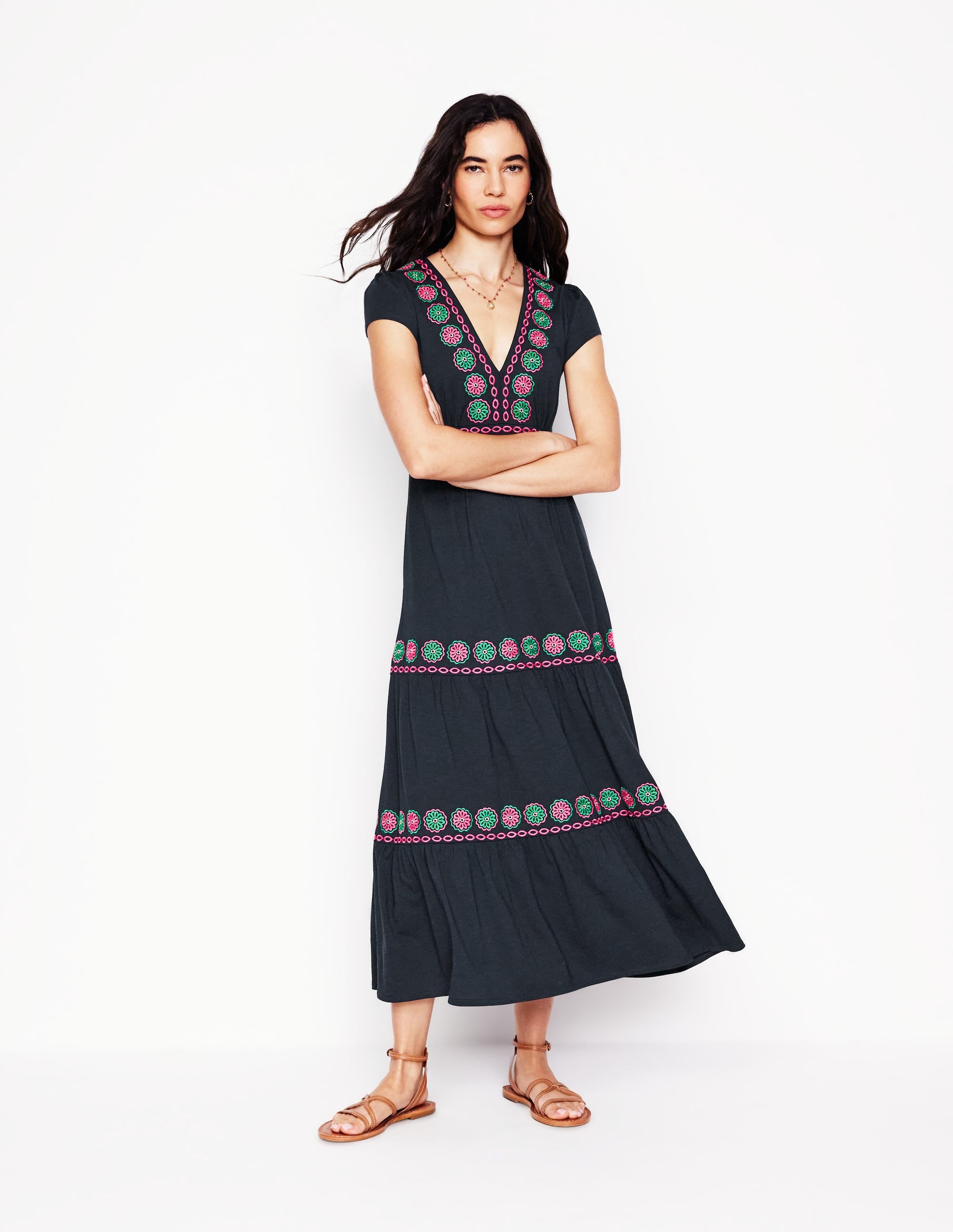 A woman wearing a navy embroidered midi dress with floral patterns, V-neckline, short sleeves, tiered design, and side seam pockets.