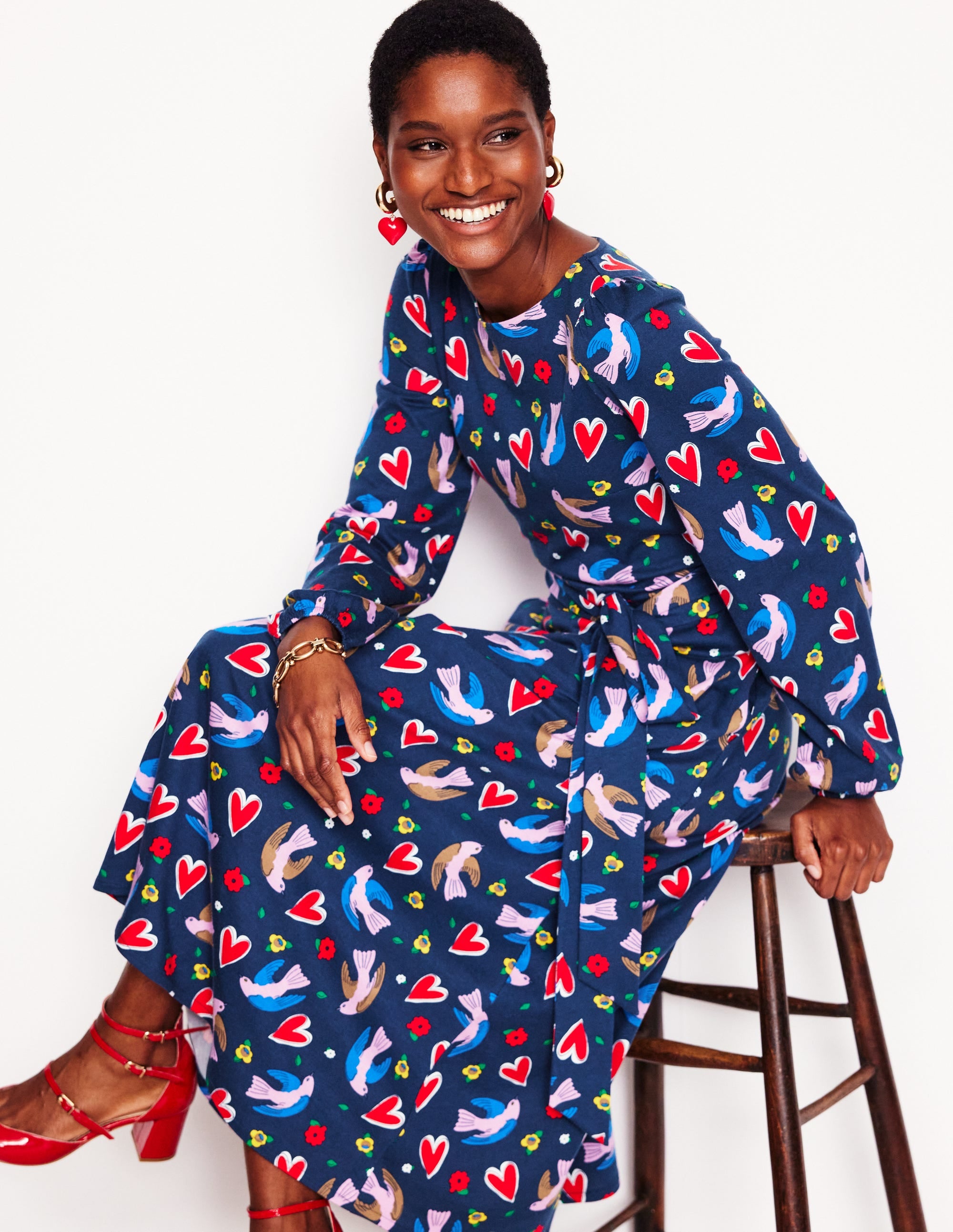A woman wearing a navy blue midi dress with a colorful heart, bird, and flower print, sitting on a wooden stool, smiling, with red shoes and heart-shaped earrings.