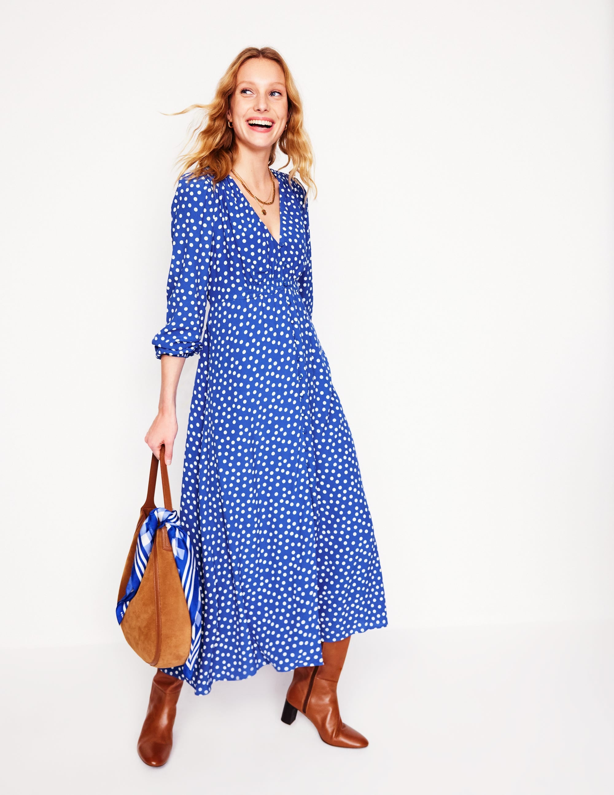 A woman wearing a bright blue tea dress with white abstract dots, button-front, tied waist, and mid-calf length, paired with brown boots and carrying a tan bag.